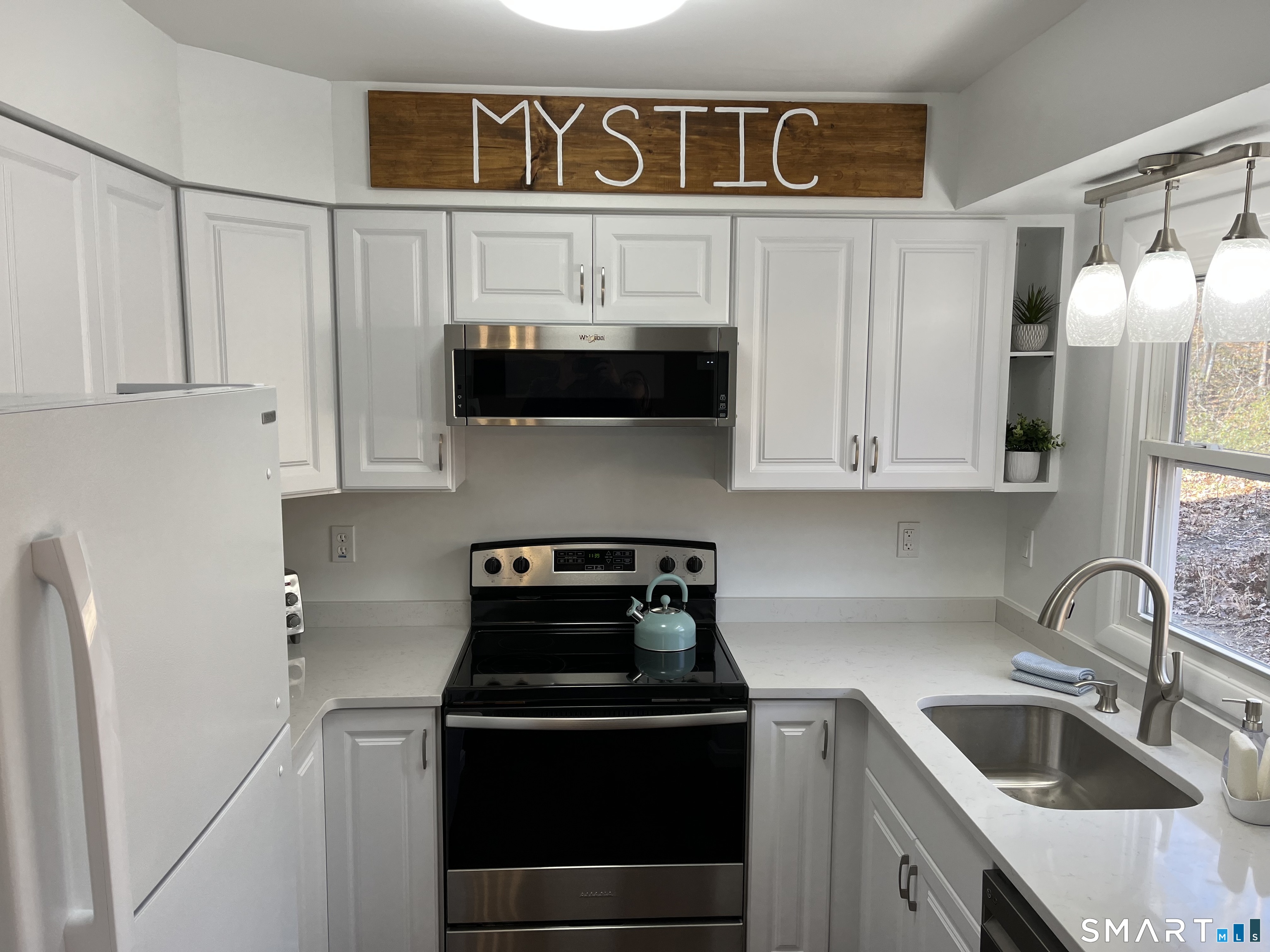 Bright white kitchen with stainless steel appliances, a black range, and a wooden “MYSTIC” sign.