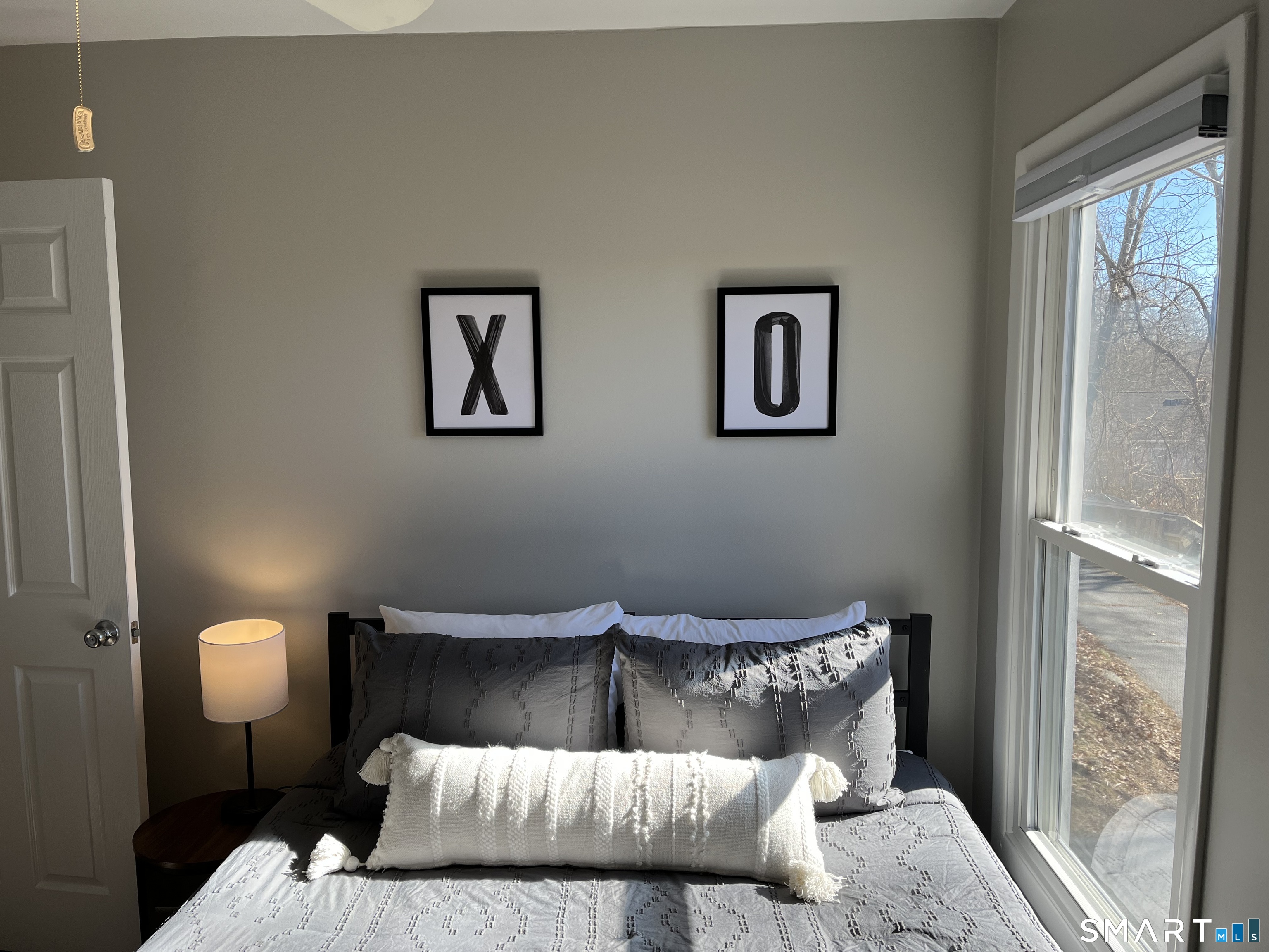 Bedroom with gray-and-white bedding, framed X and O prints, and a large sunlit window.