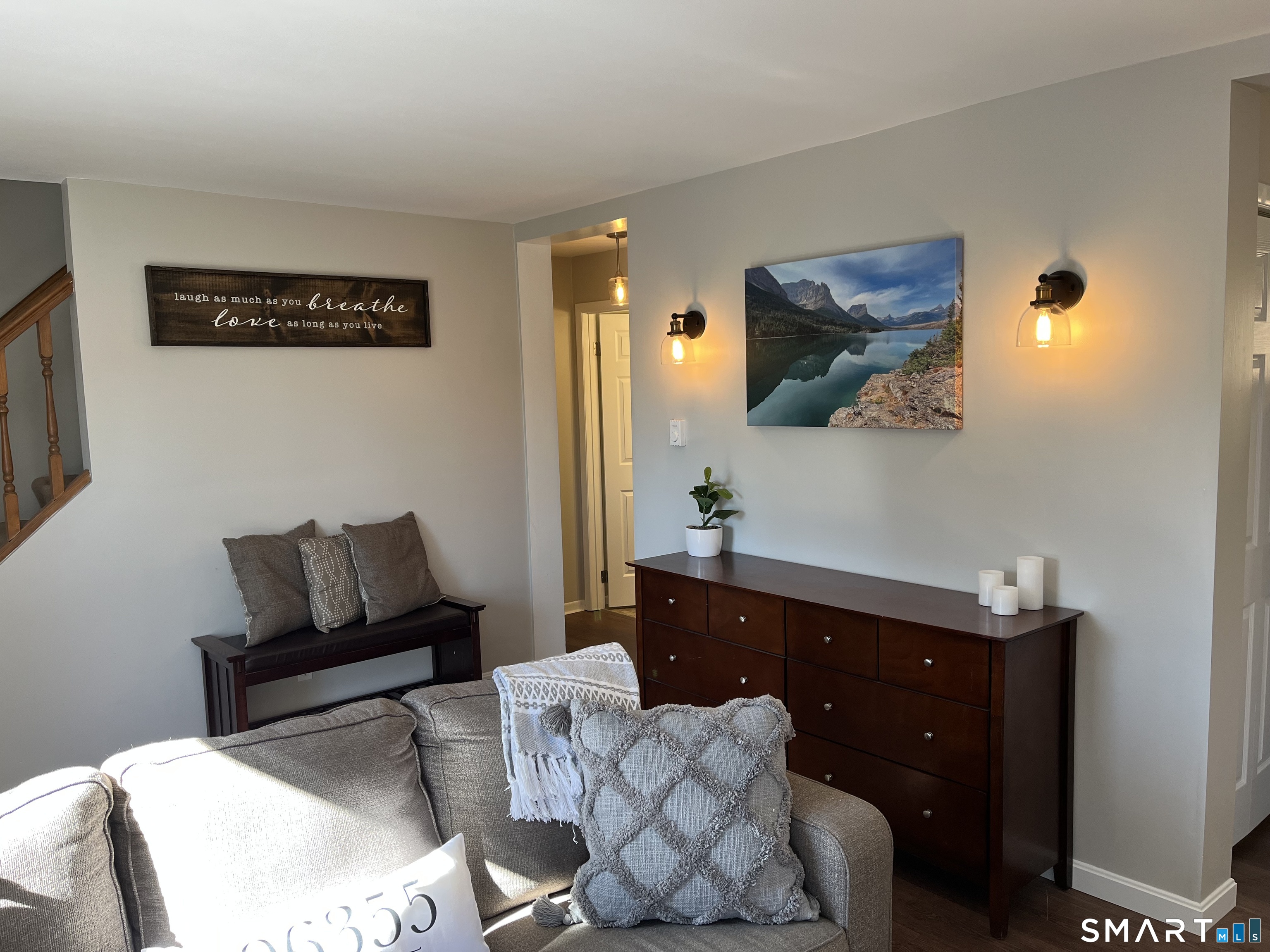 Living room with a gray sofa, dark wood dresser, warm sconces, and framed mountain-lake photograph.