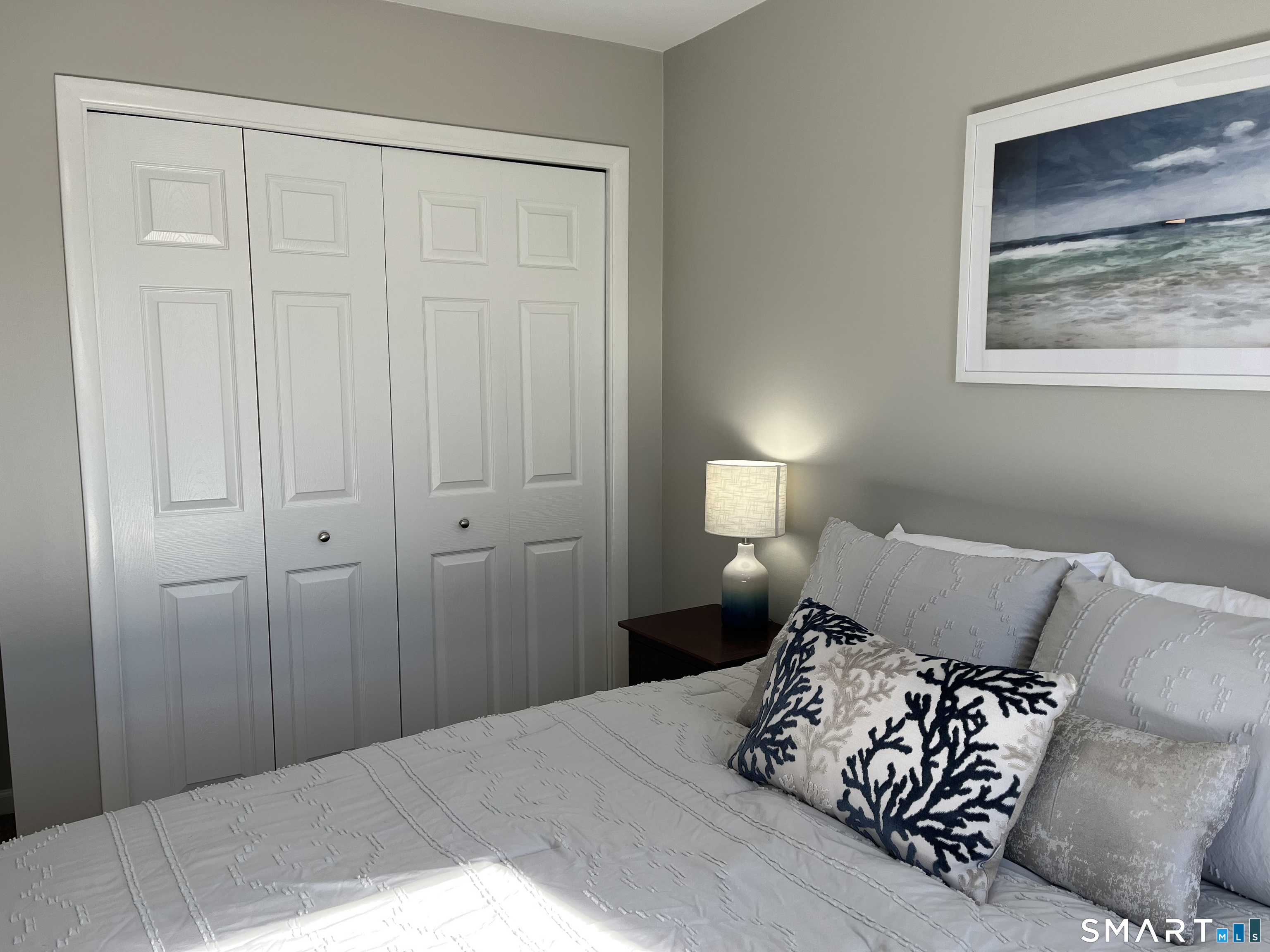 Furnished bedroom with white paneled closet doors, a bedside lamp, and framed ocean artwork.