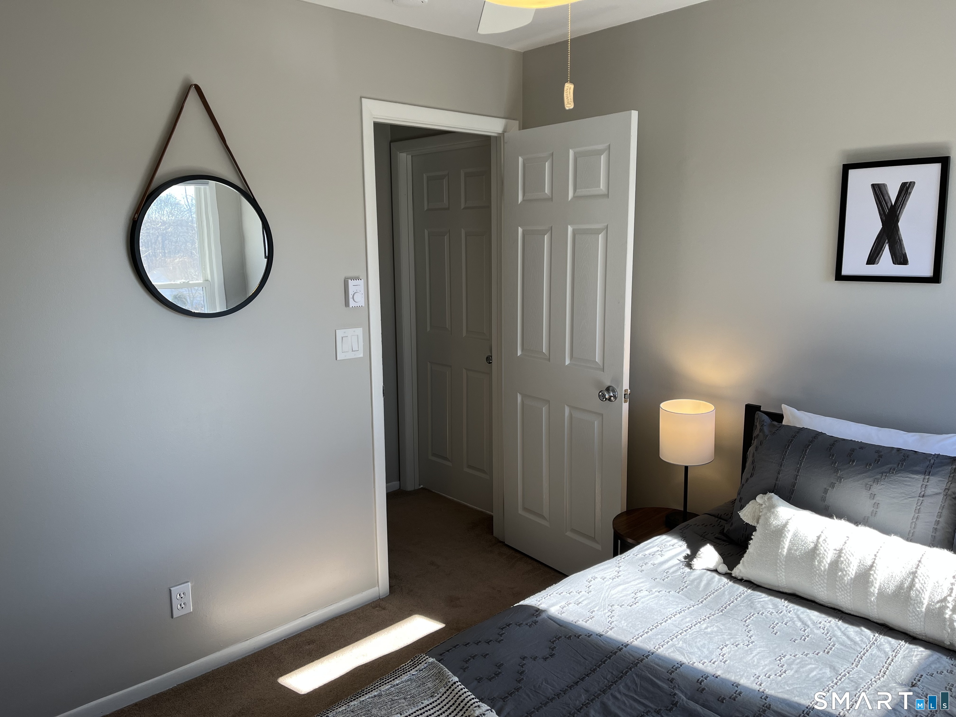 Bedroom with a gray-and-white bed, round hanging mirror, bedside lamp, and open doorway.