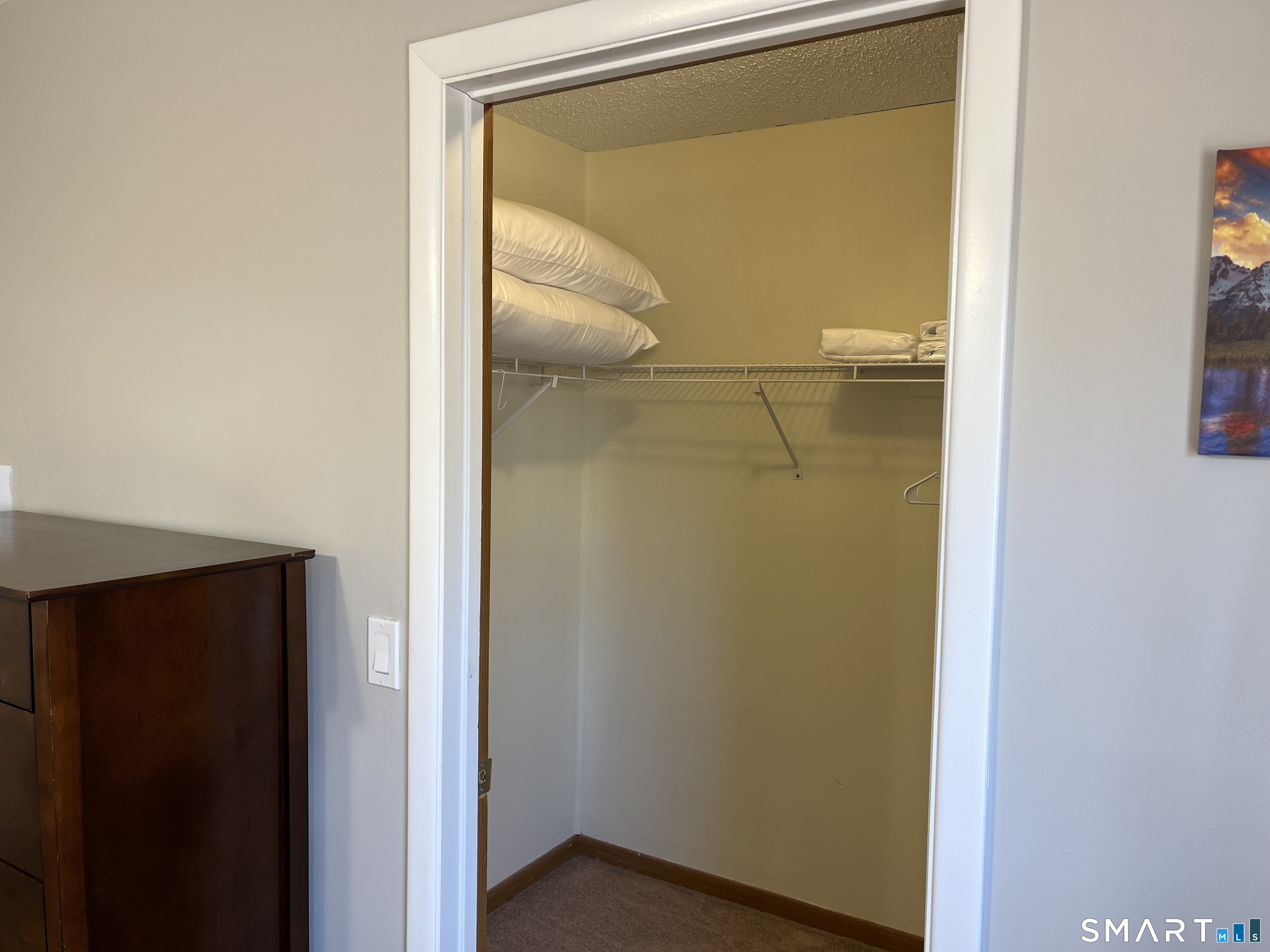 Open walk-in closet with a wire shelf holding pillows and a dark wooden dresser beside the doorway.