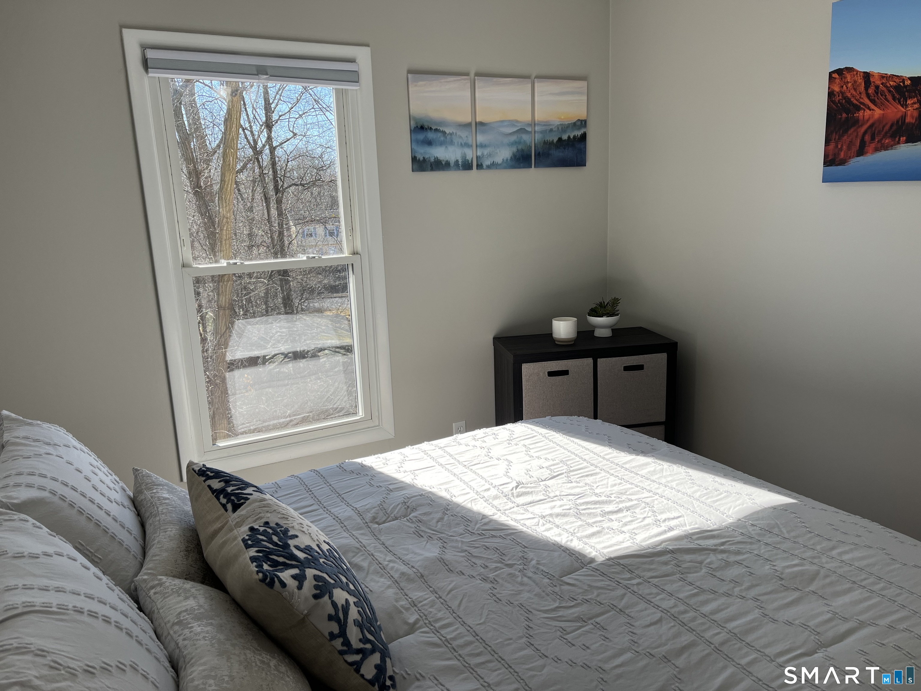 Staged bedroom with a white quilted bed, large window, and sunlight across the bedding.
