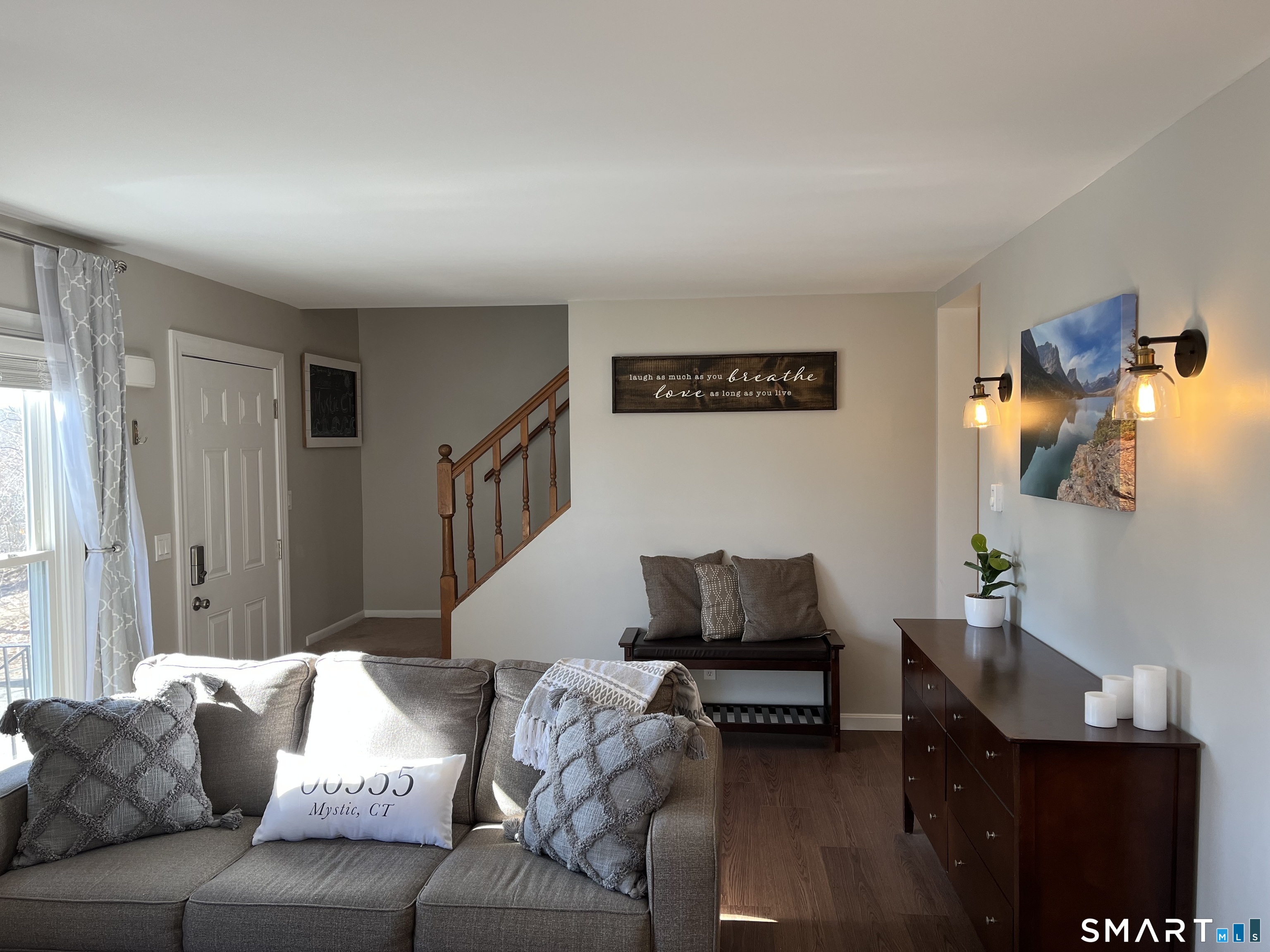 Furnished living room with a gray sectional, sunlit window, and wooden staircase railing.