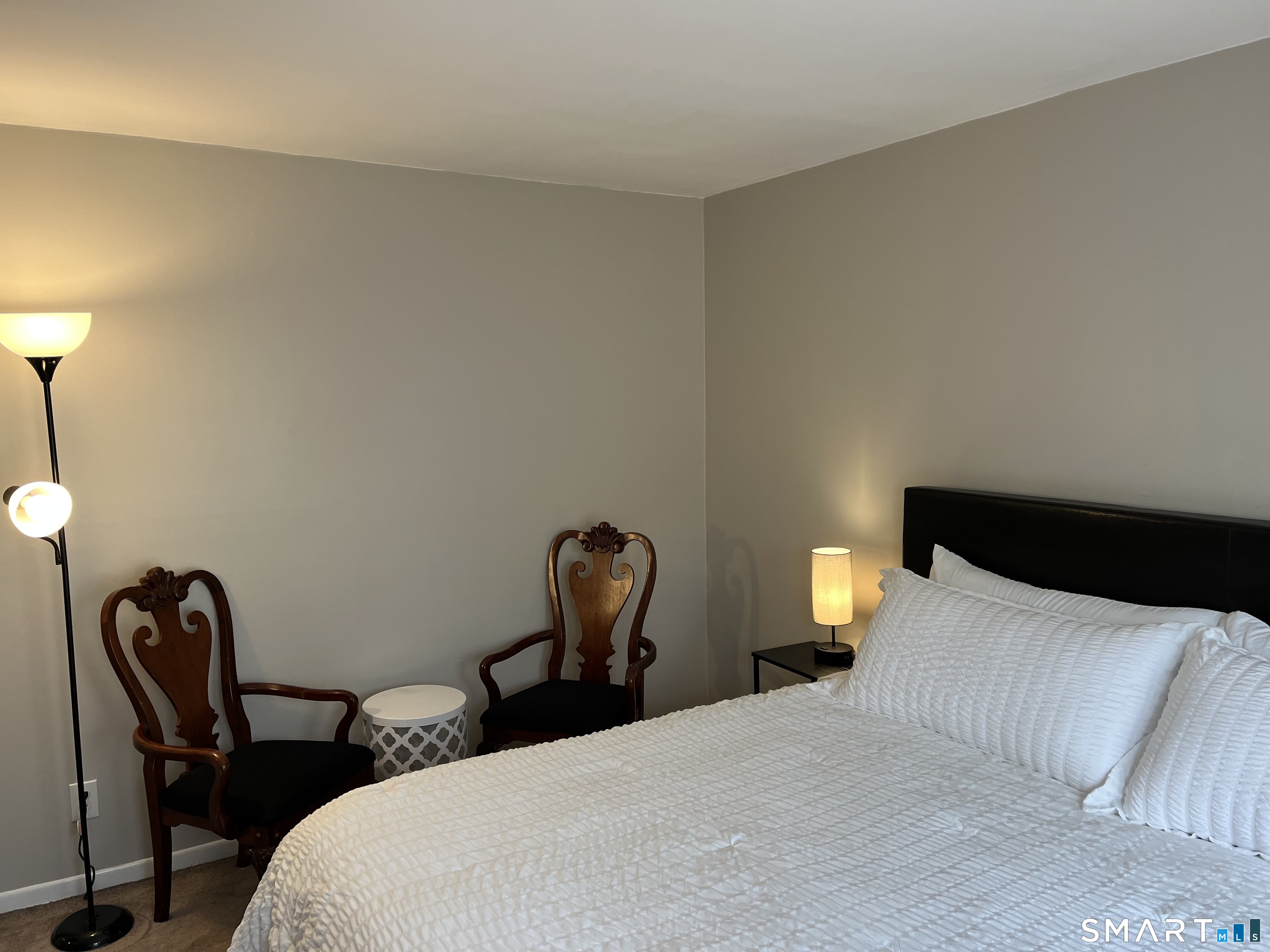Furnished bedroom with white bedding, a dark headboard, and carved wooden chairs.