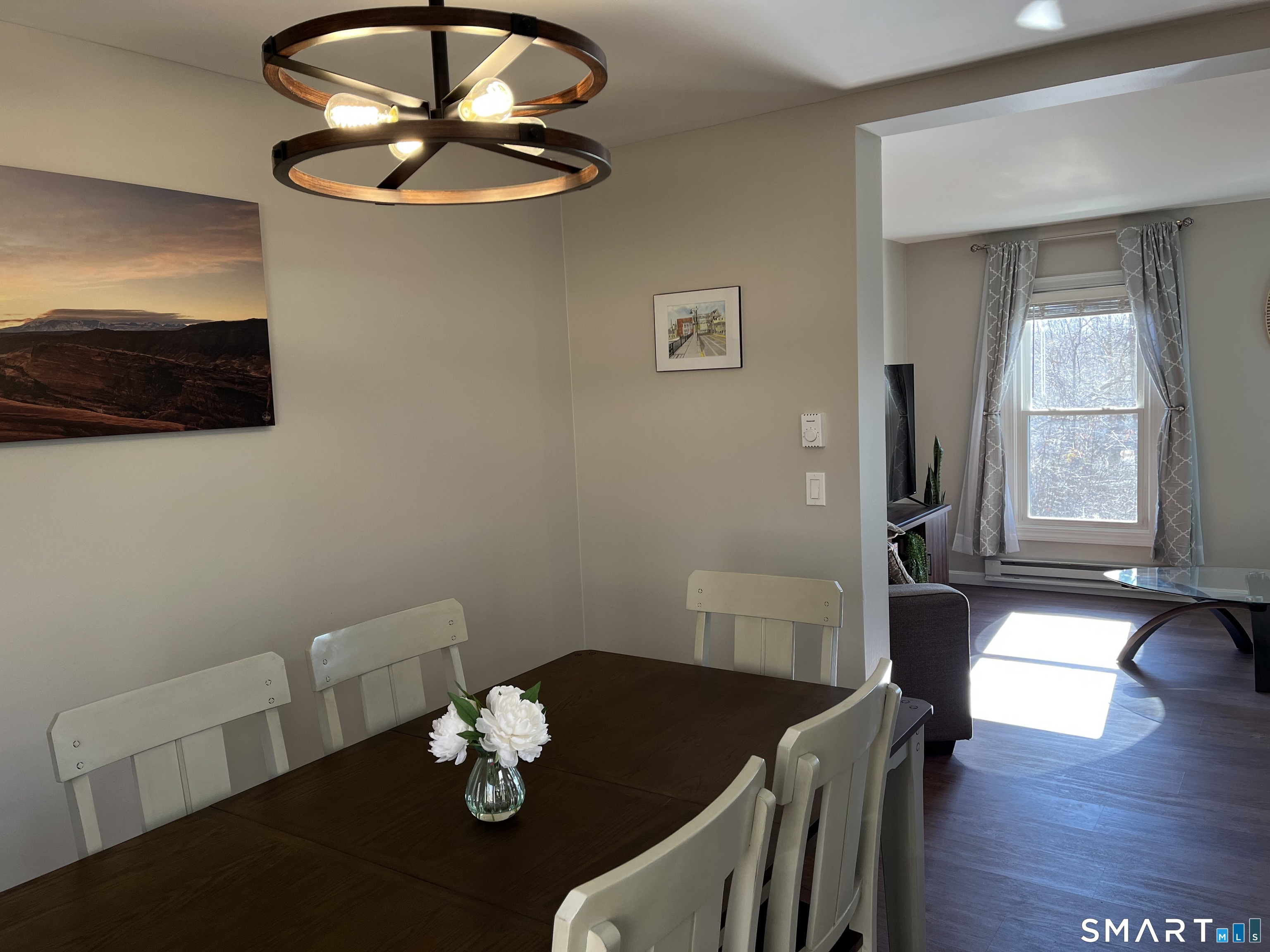 Dining room with a dark wood table, white chairs, and a circular pendant light beside a bright sitting area.