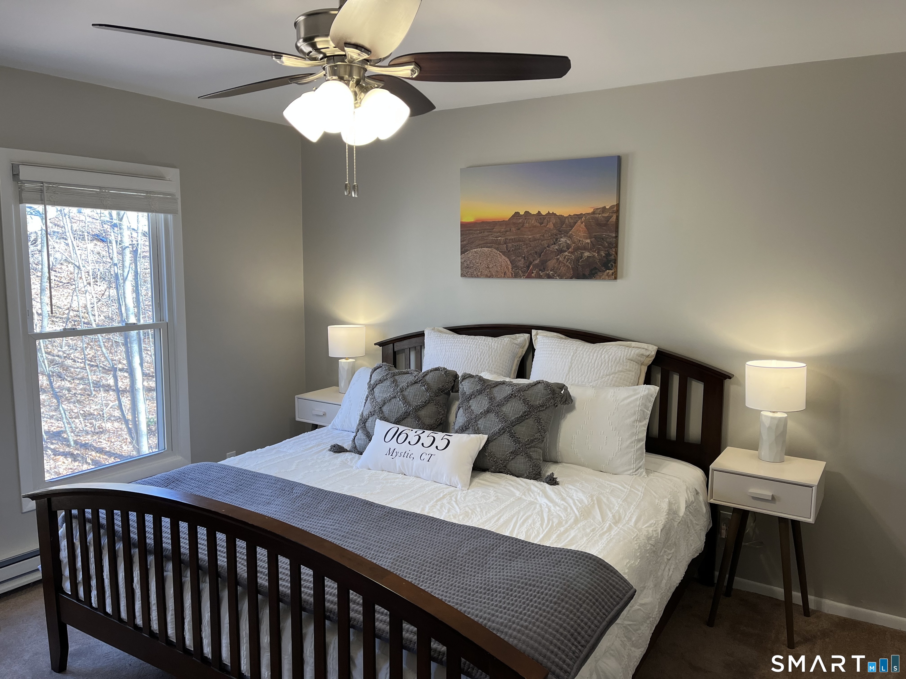 Furnished bedroom with a dark wood bed, layered white and gray bedding, and a large window overlooking trees.