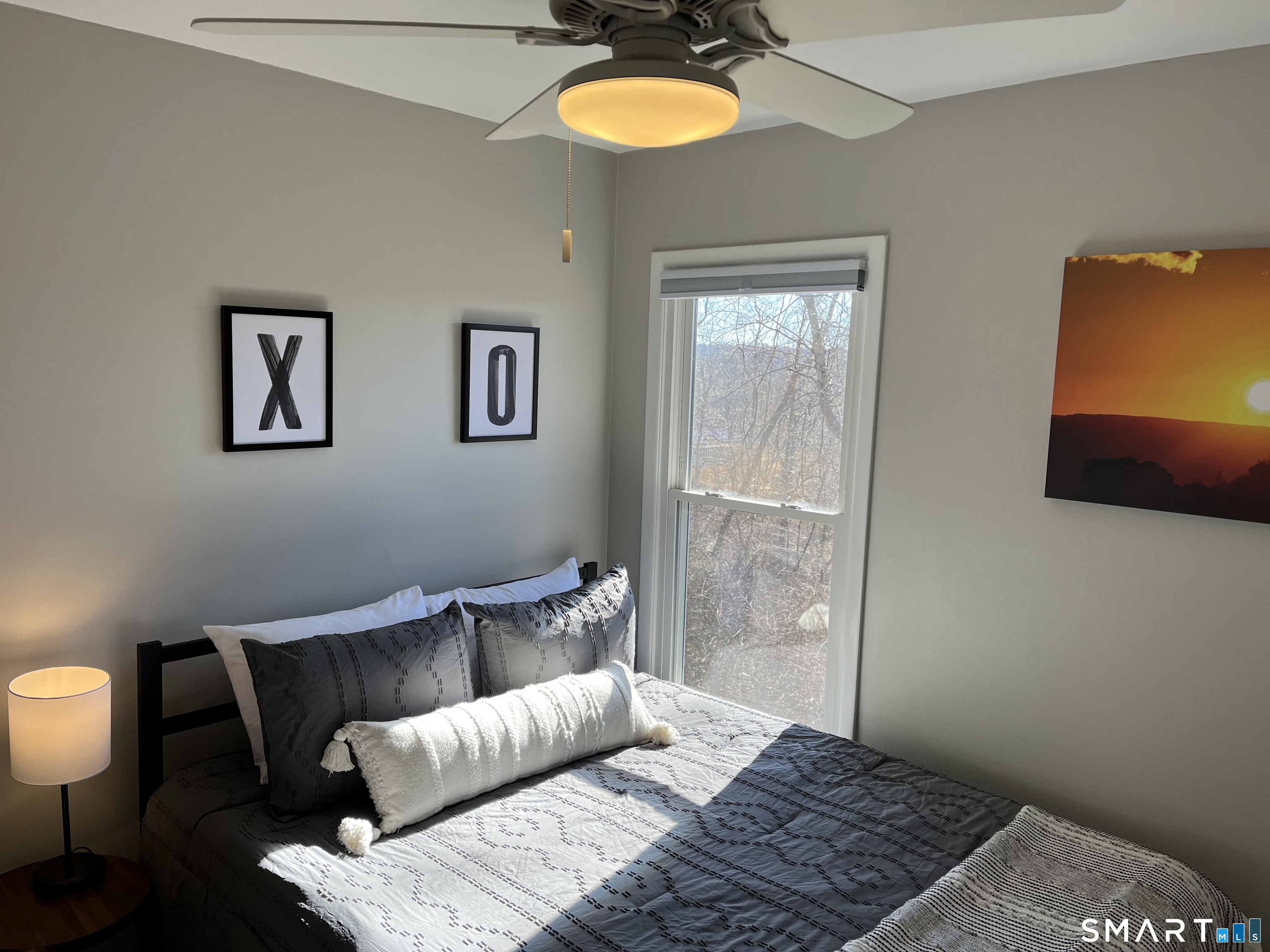 Staged bedroom with gray-and-white bedding, framed X and O prints, and a sunlit window.