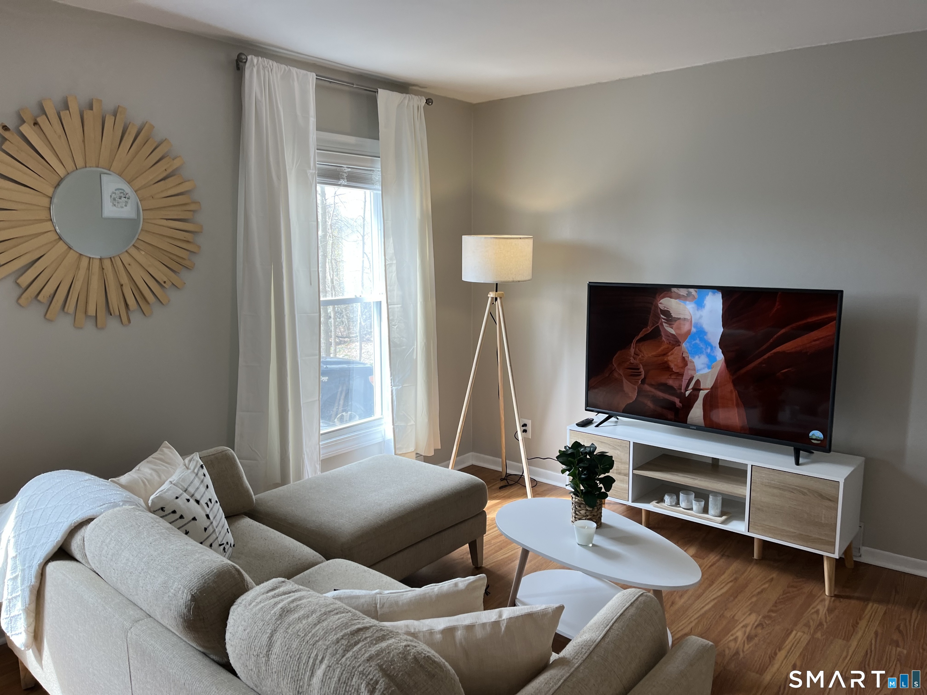 Staged living room with a light sectional, oval coffee table, and TV beside a bright curtained window.