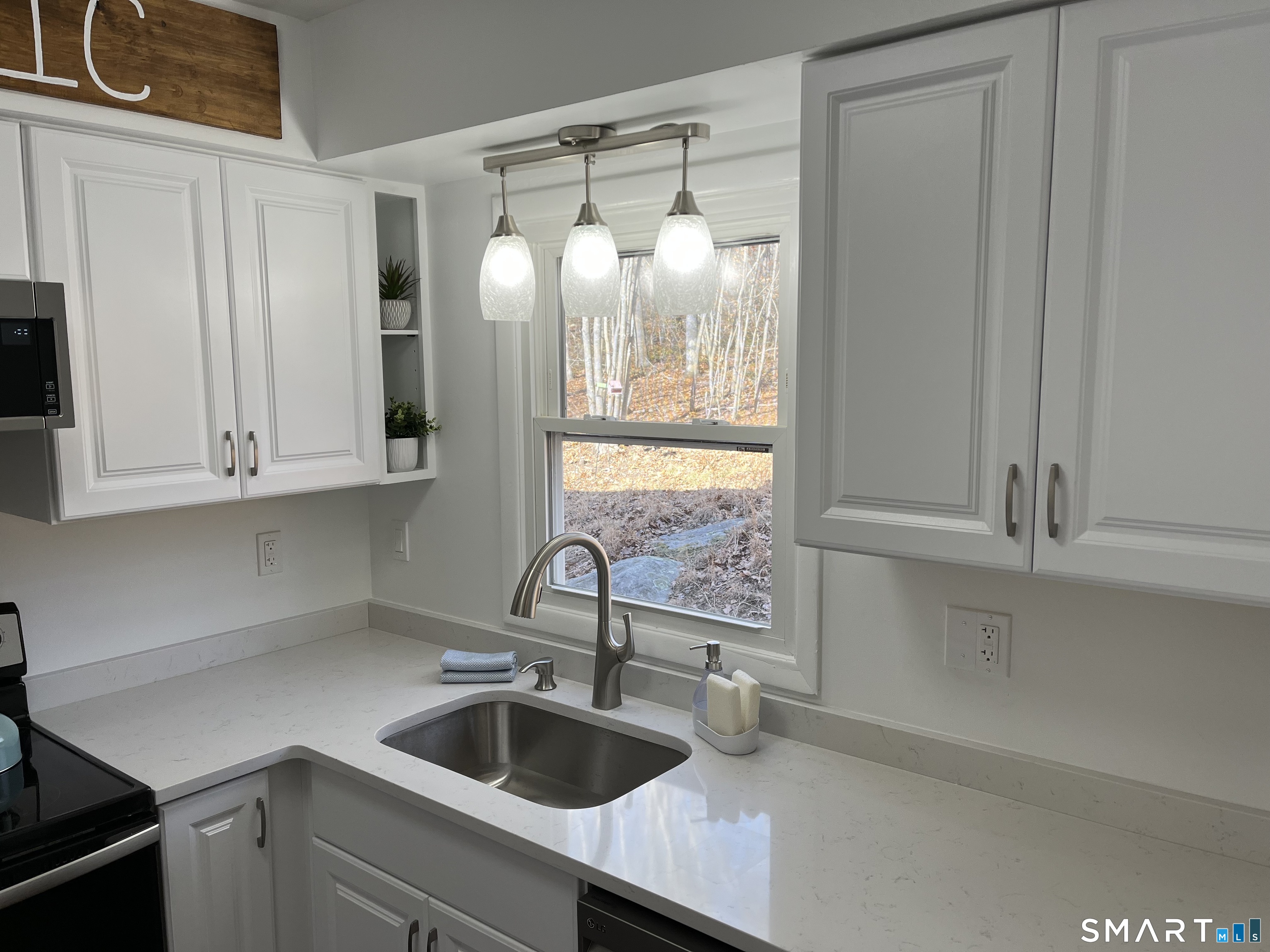 Bright kitchen with white cabinets, pale stone counters, and a sink beneath a window overlooking autumn trees.
