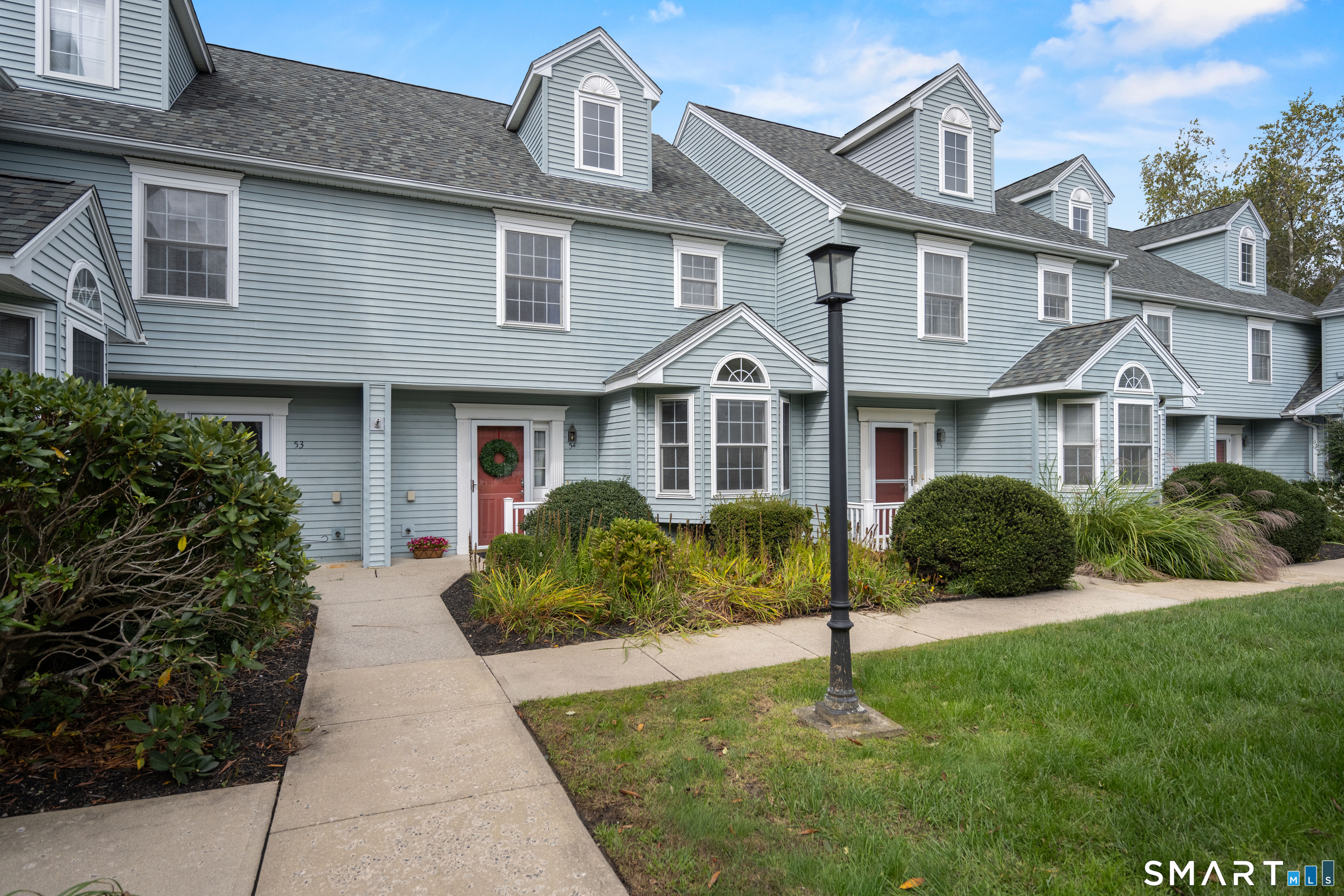 Light blue townhouse exterior with red entry doors, landscaped shrubs, and a sidewalk.