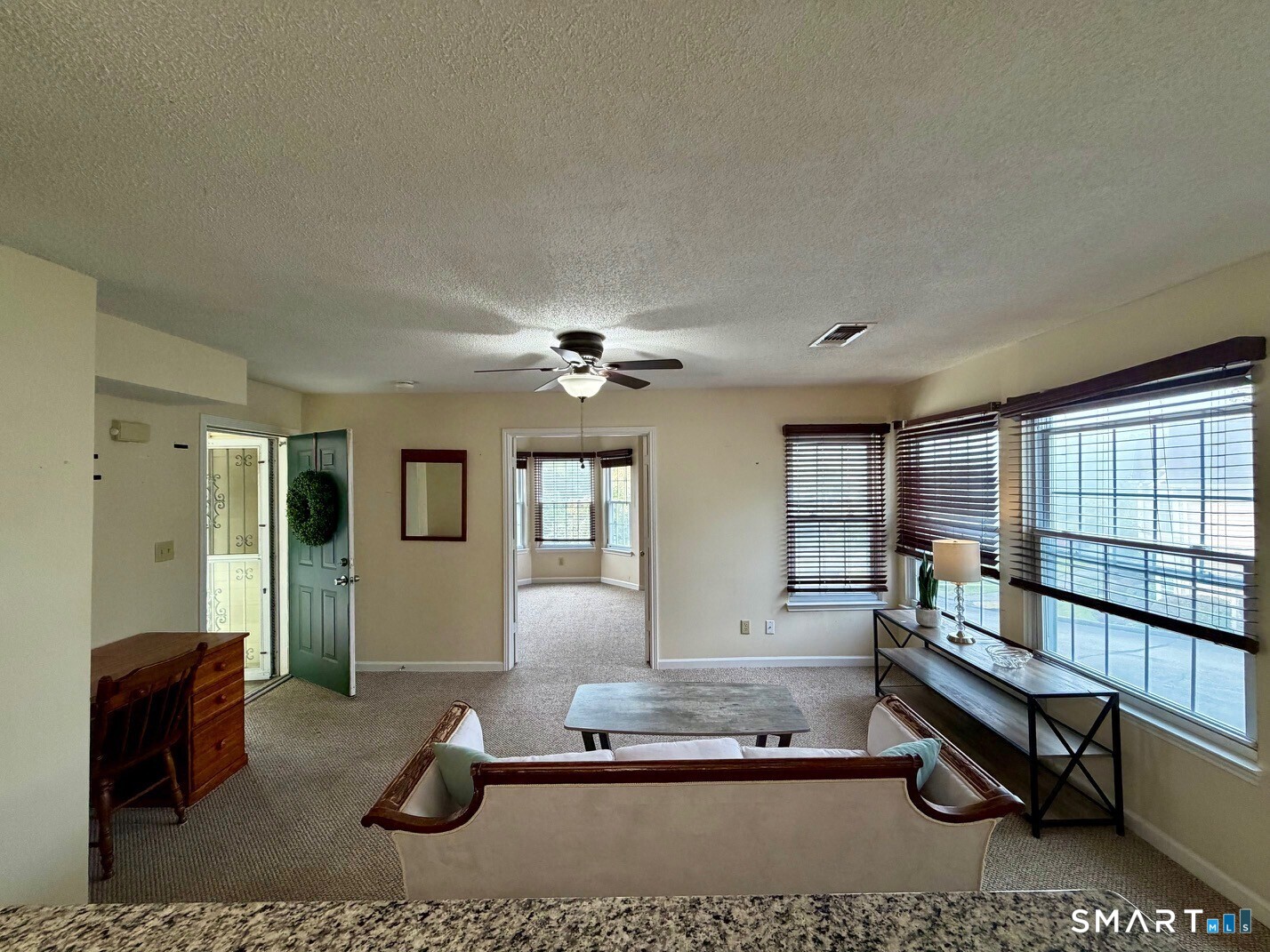 Living room with wide blinded windows, carpet, ceiling fan, and a doorway to a bright adjoining room.