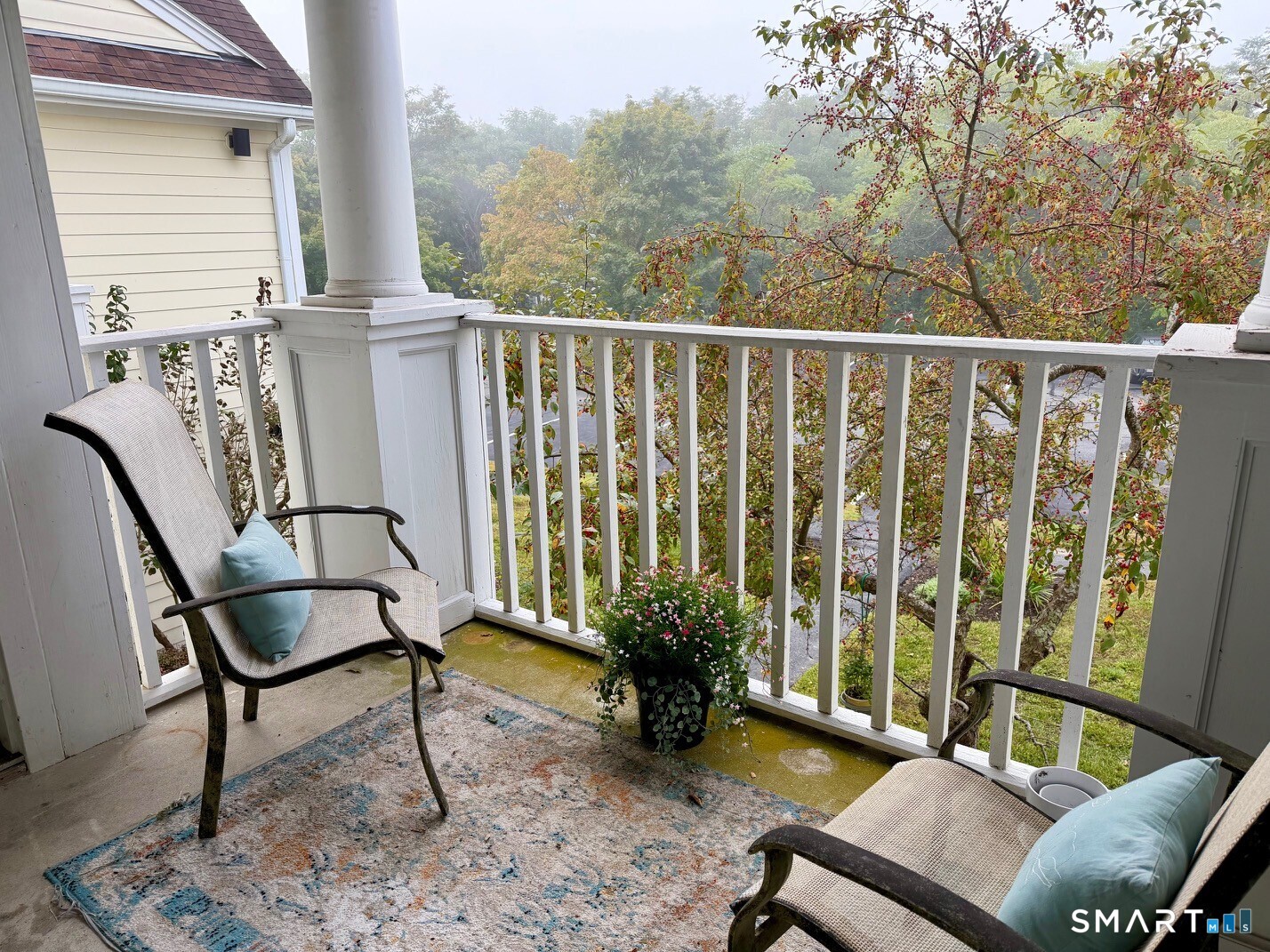 Furnished porch with white railings, two chairs, and a view of autumn trees.