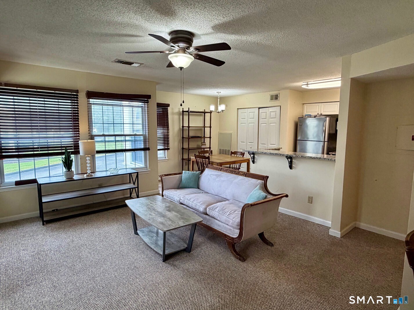 Living room with large windows, a sofa, ceiling fan, and adjoining dining area and kitchen.