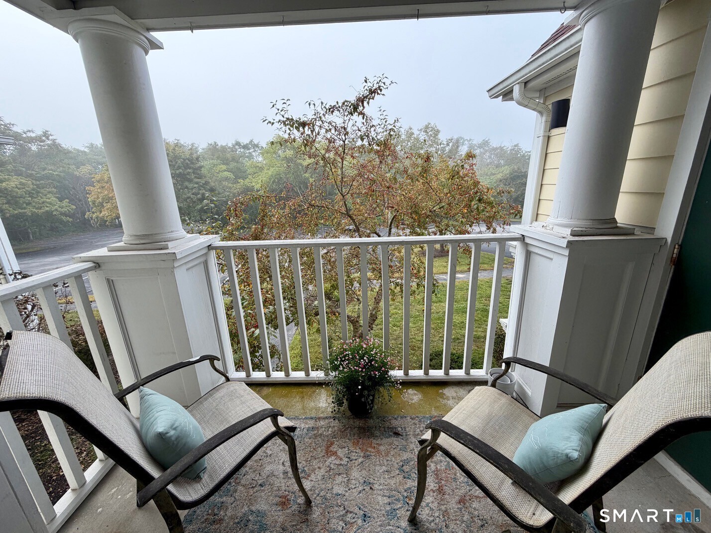 Covered porch with white columns, cushioned chairs, and a view of autumn trees and parking area.