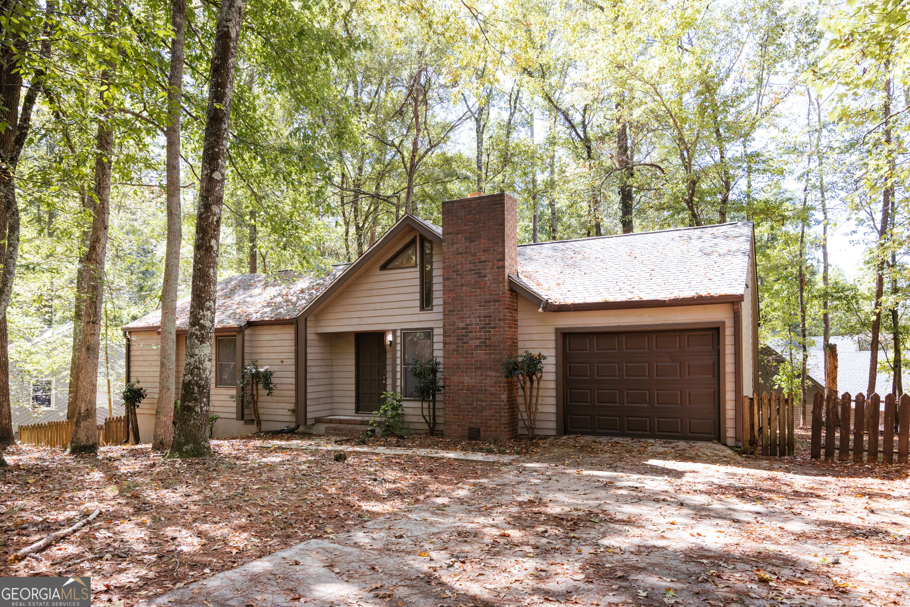 Beige-sided house with a brick chimney and attached garage surrounded by mature trees.