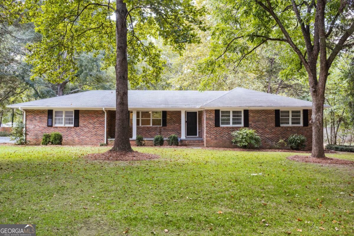 Single-story brick ranch house with black shutters, framed by mature trees across a broad lawn.