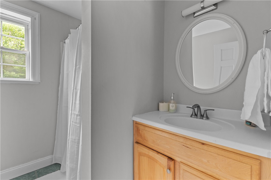Bathroom with a light wood vanity, oval mirror, and shower curtain beside a window.