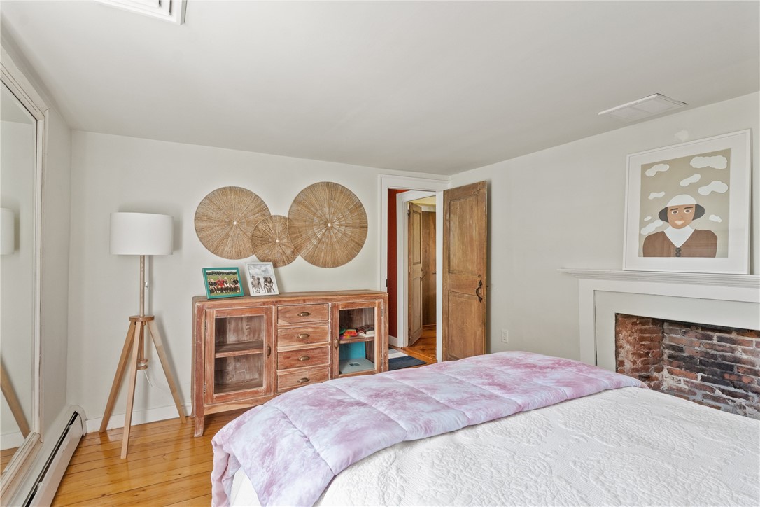 Bedroom with a pale pink quilt, wooden dresser, hardwood floors, and brick fireplace.