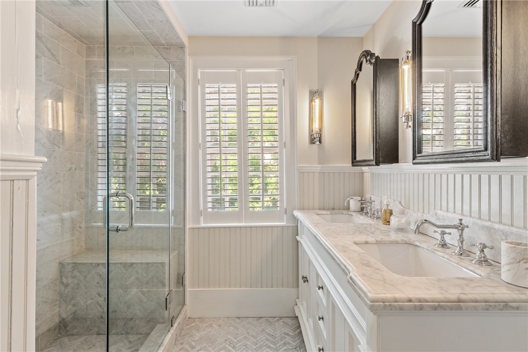 Bathroom with a marble glass-enclosed shower, double vanity, and plantation shutters.