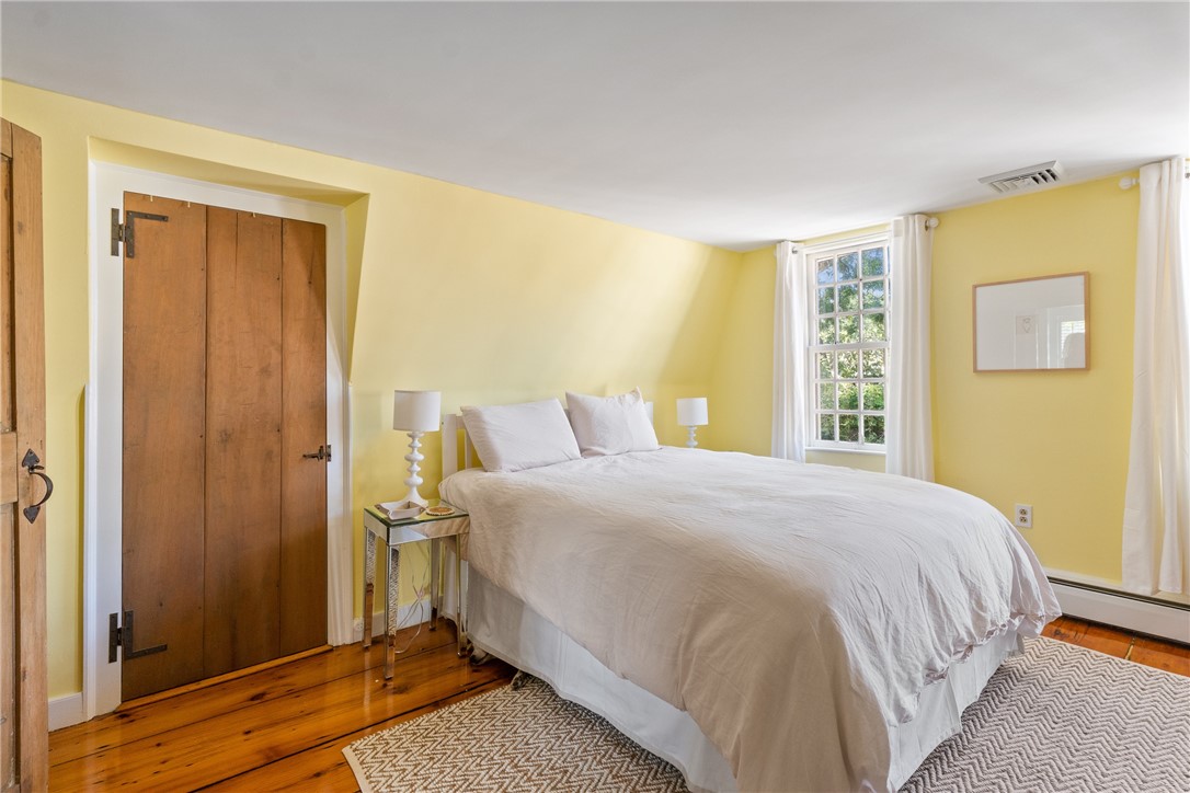 Bedroom with a white bed, yellow walls, polished wood floors, and a tall window beside a rustic wooden door.