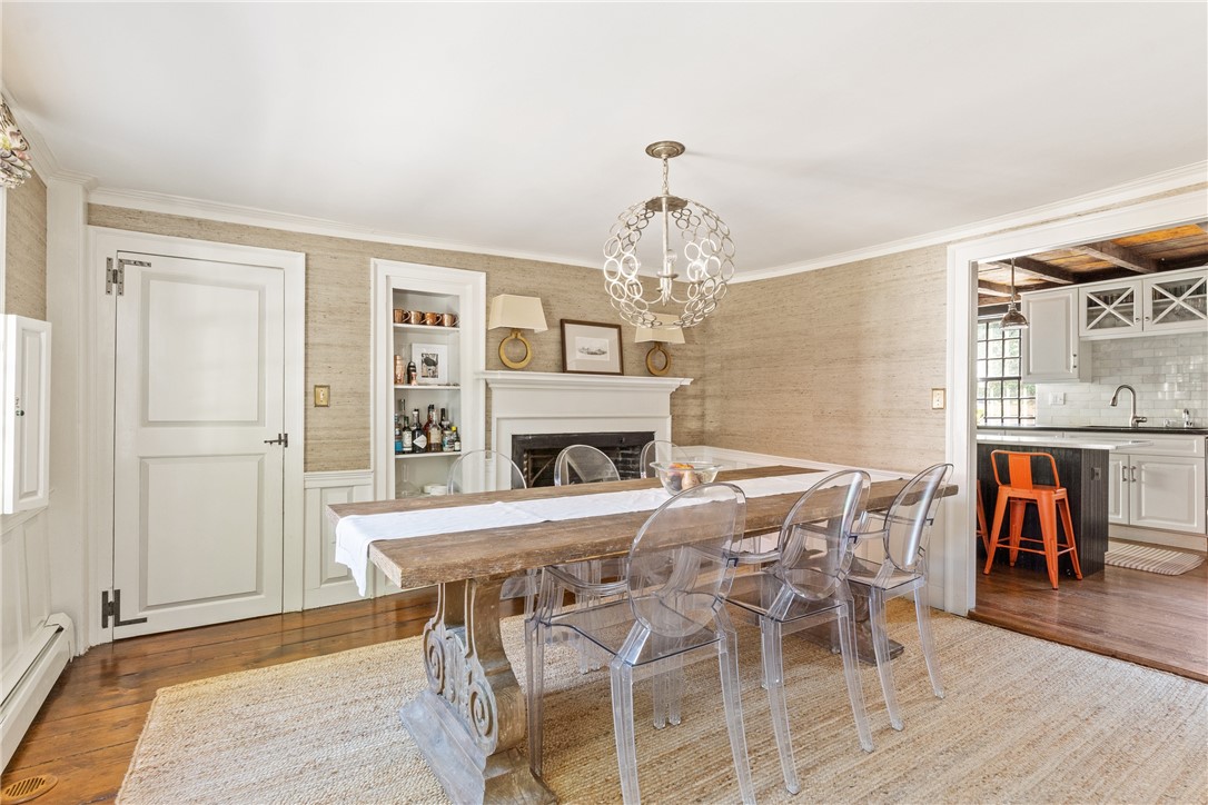 Dining room with a rustic wood table, clear chairs, white fireplace, and ornate chandelier beside an open kitchen.
