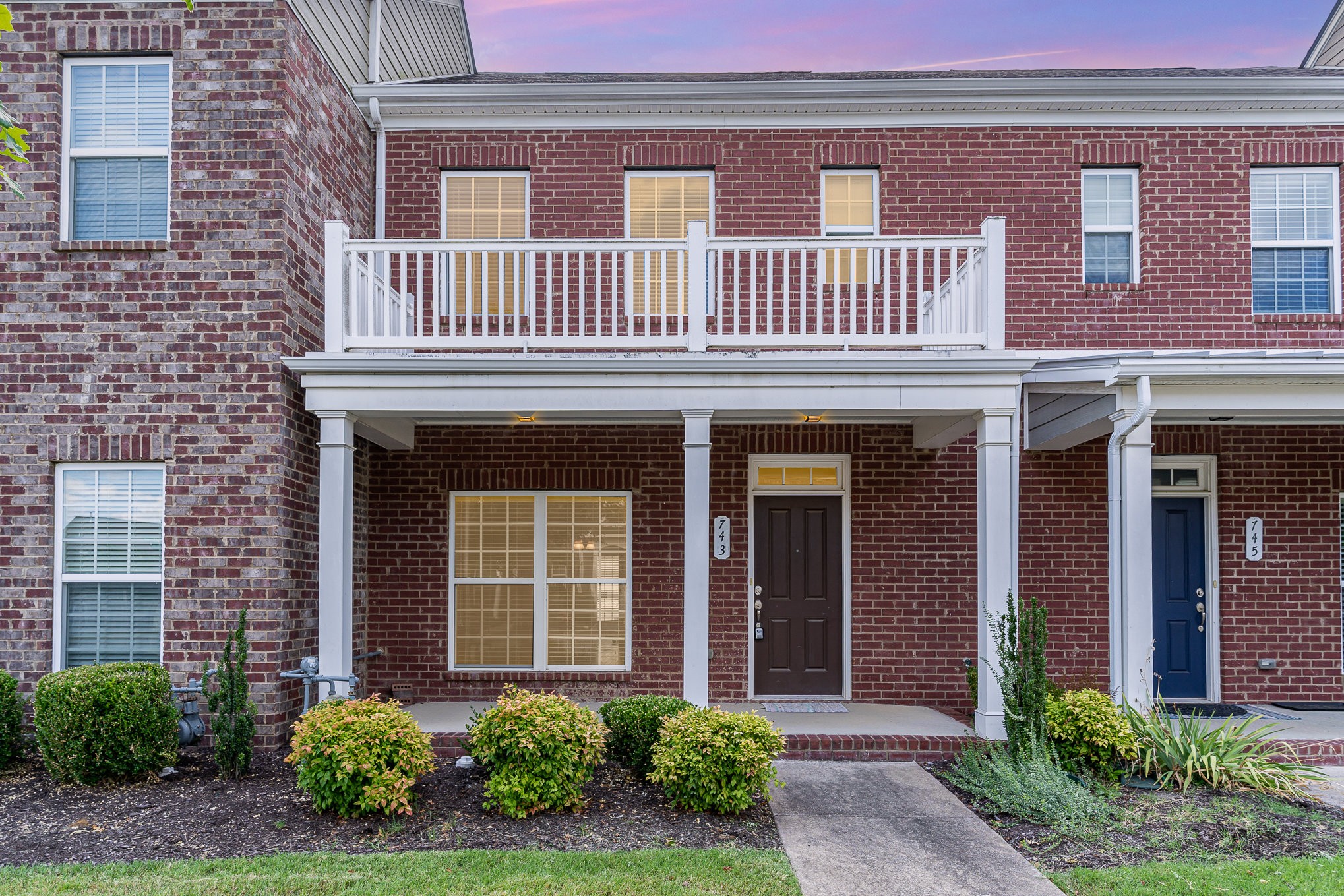 Brick townhouse with a white-railed balcony, covered porch, and landscaped front walkway.