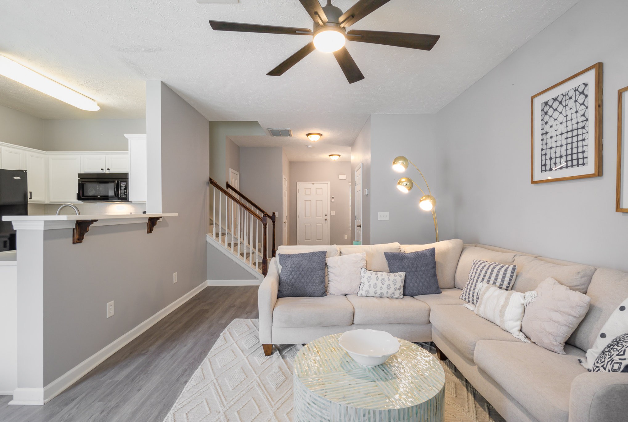 Staged living room with a light sectional, glass coffee table, and open kitchen beside the staircase.
