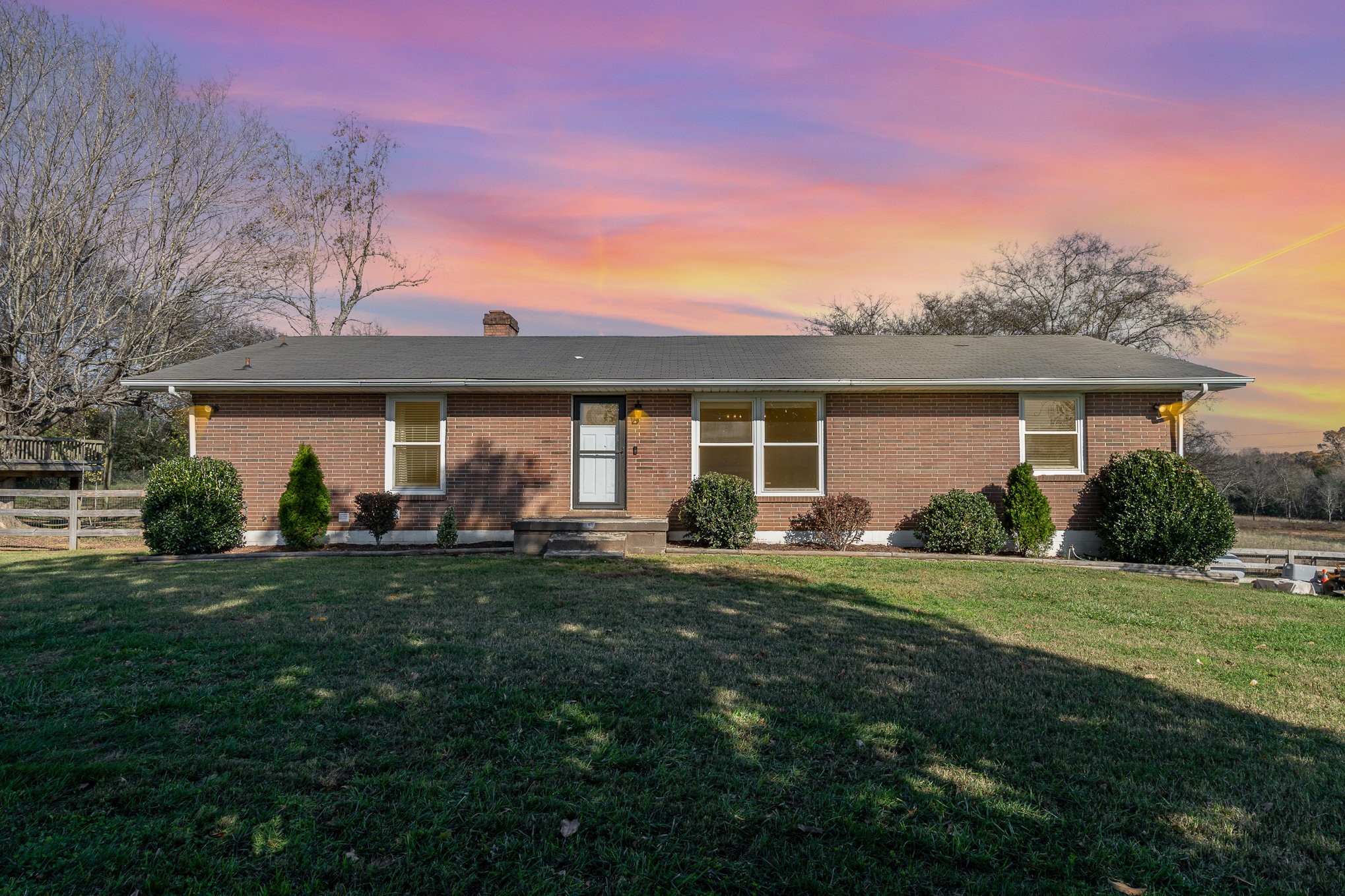 Single-story brick ranch house with trimmed shrubs beneath a vivid pink and orange sunset sky.