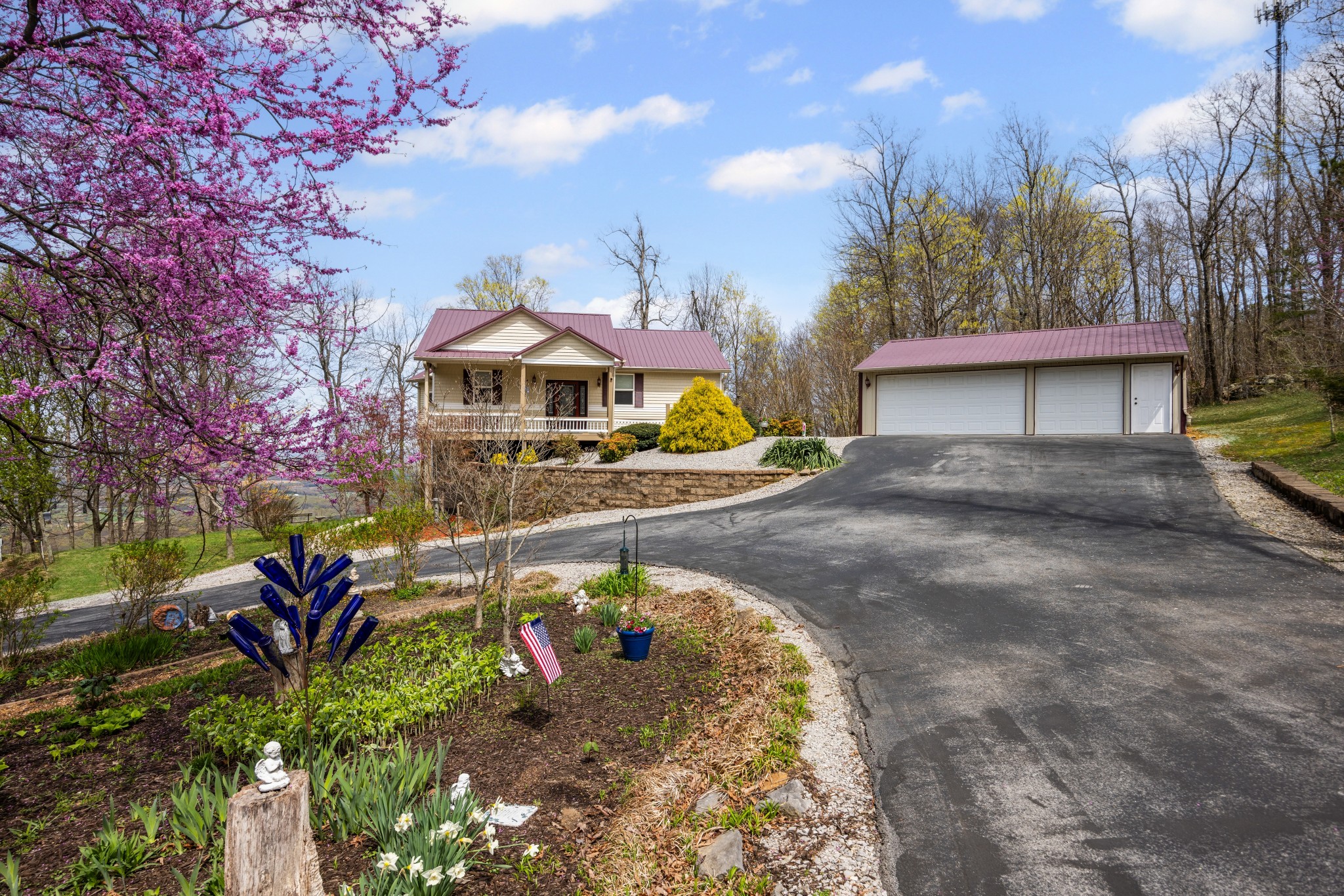 Beige home and detached two-bay garage with matching burgundy roofs, curved driveway, and blooming pink tree.