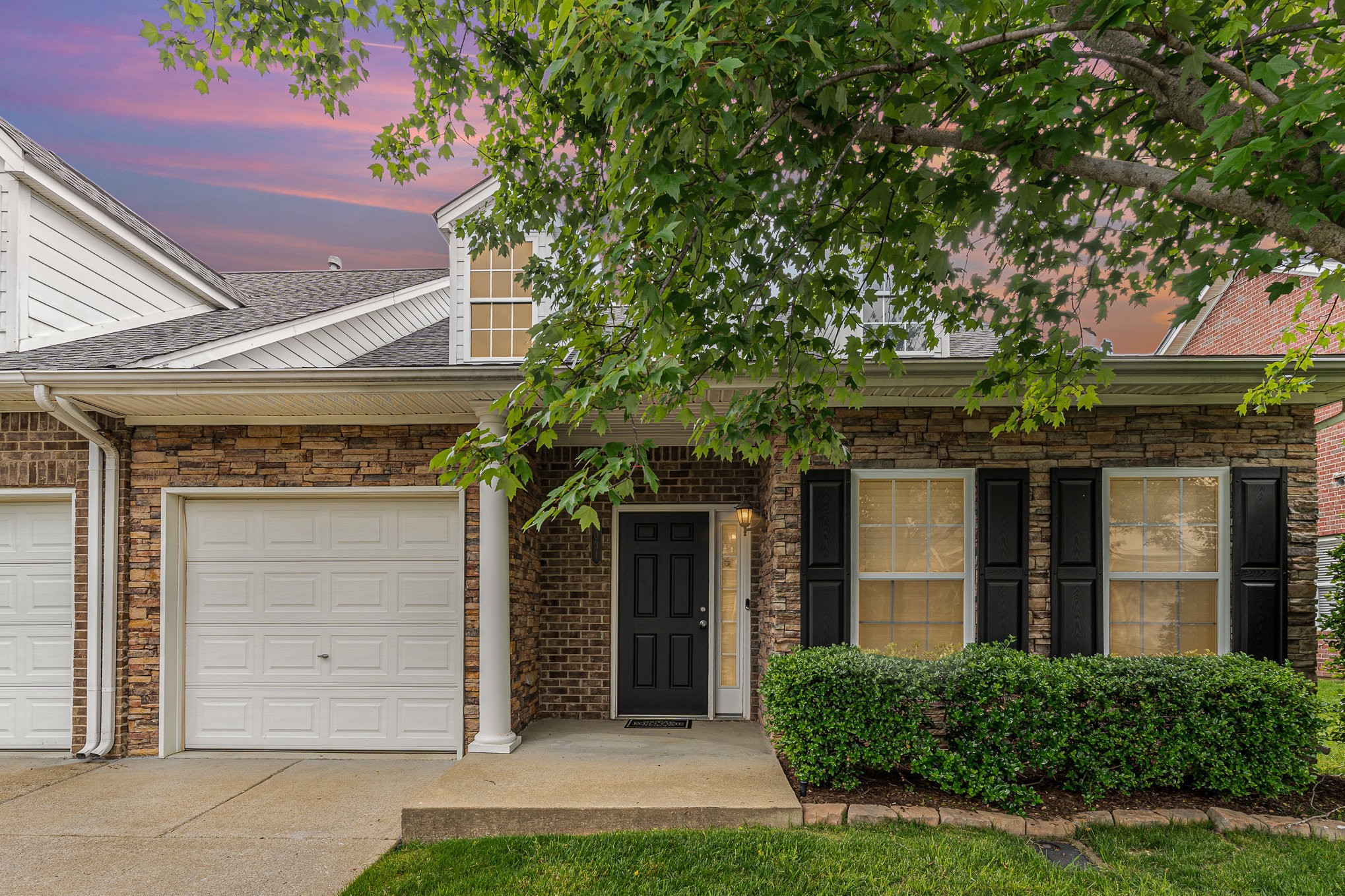 Stone-and-brick home with a white garage, black shutters, and a leafy tree at dusk.