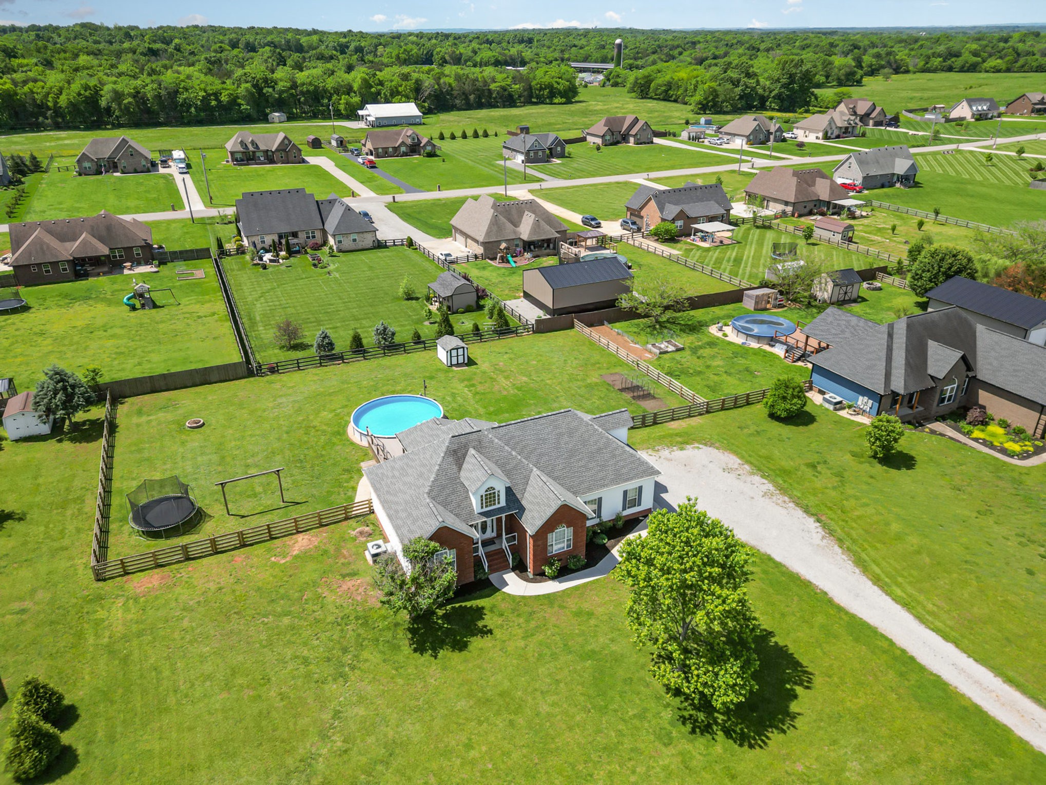 Aerial view of a home with a large fenced lawn, above-ground pool, trampoline, and outbuildings.