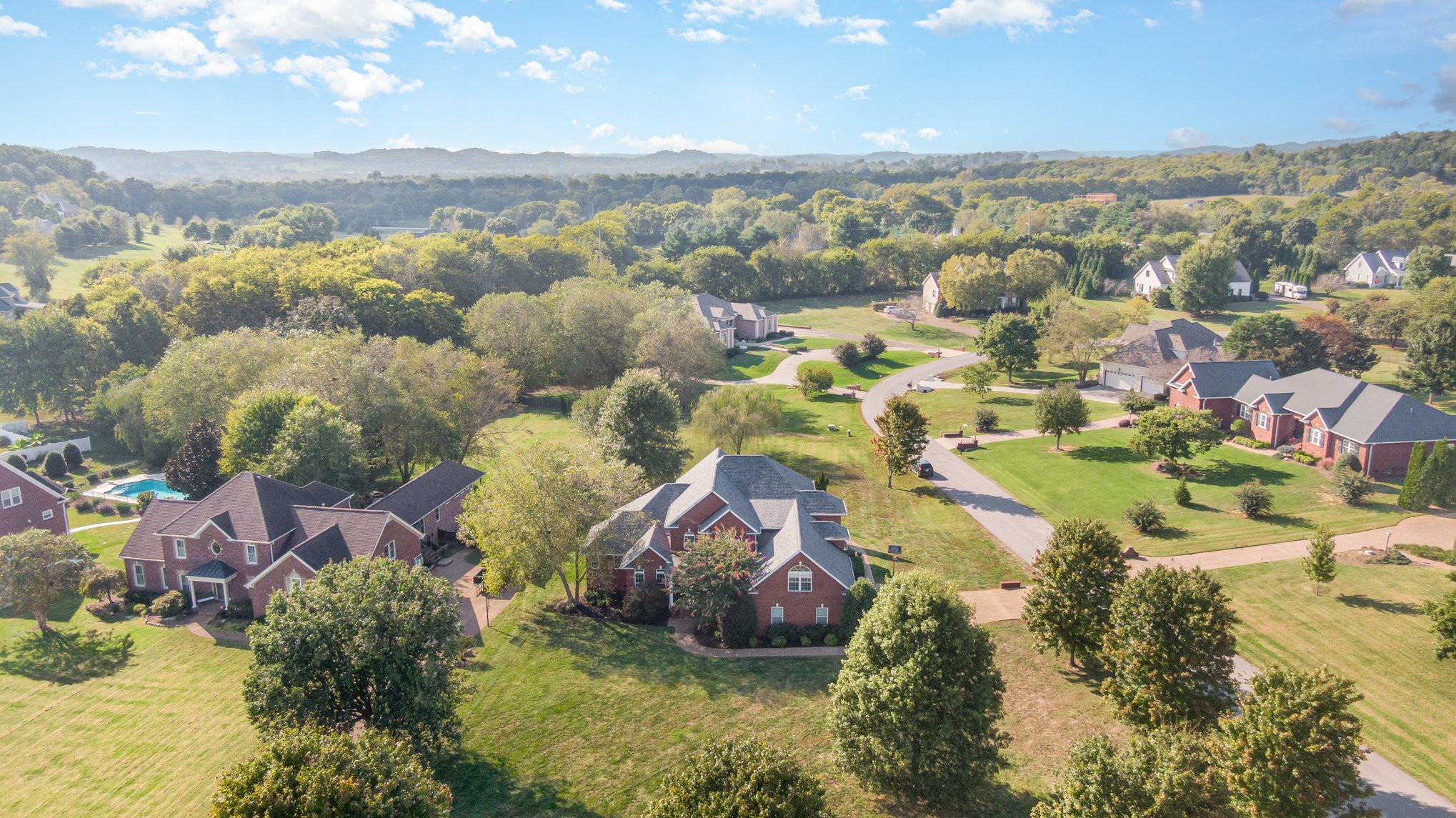 Aerial view of a leafy neighborhood with brick homes, a winding street, and distant wooded hills.
