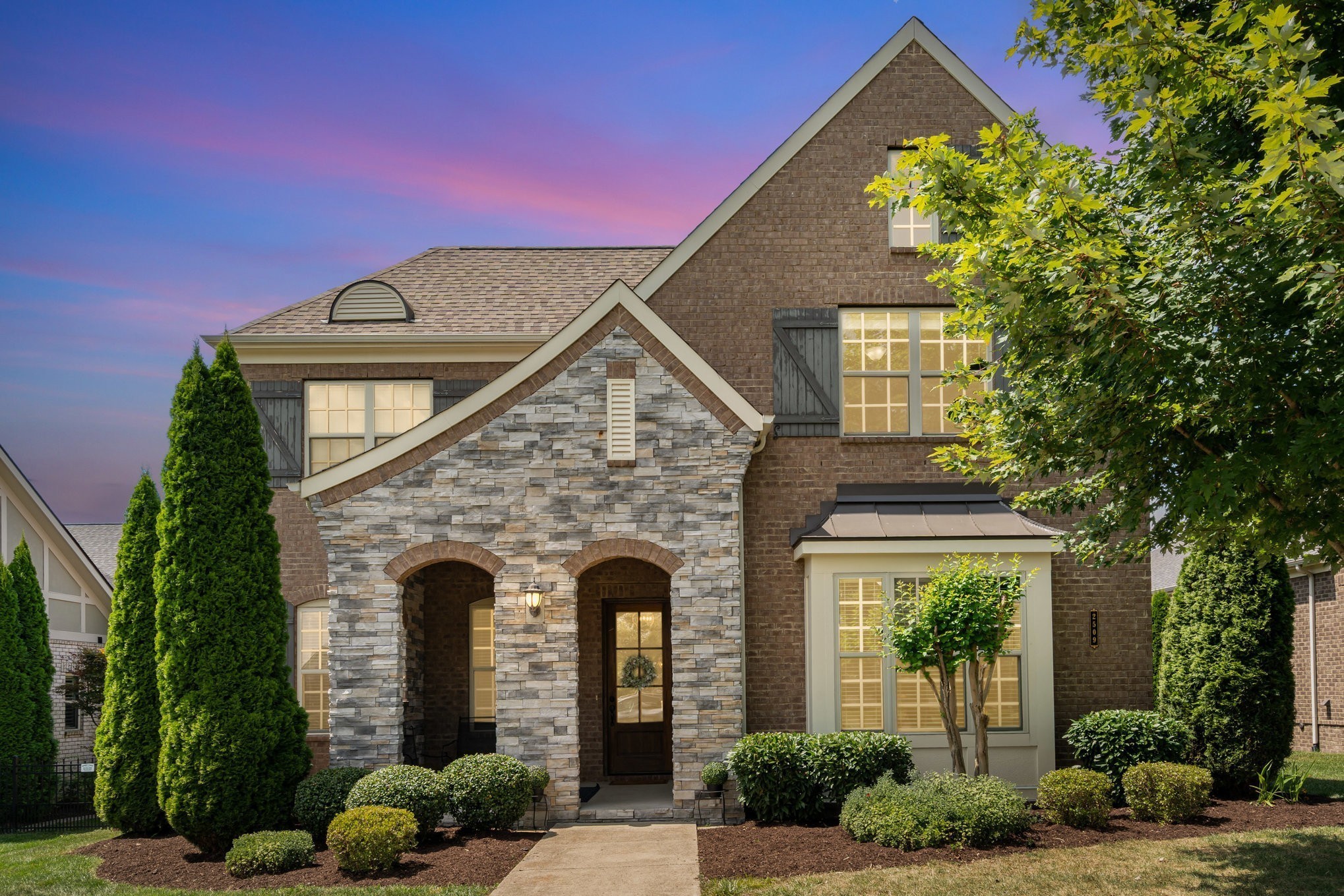 Two-story stone-and-brick home with an arched entry and landscaped front yard at dusk.