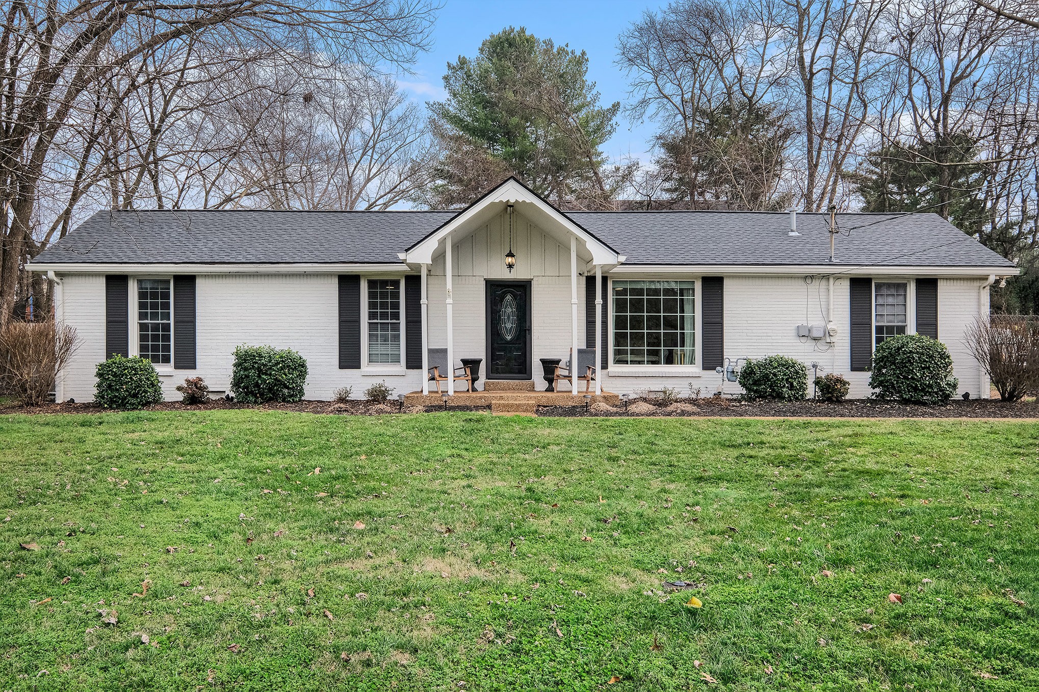 White brick ranch house with black shutters and a covered entry, viewed across a green lawn.