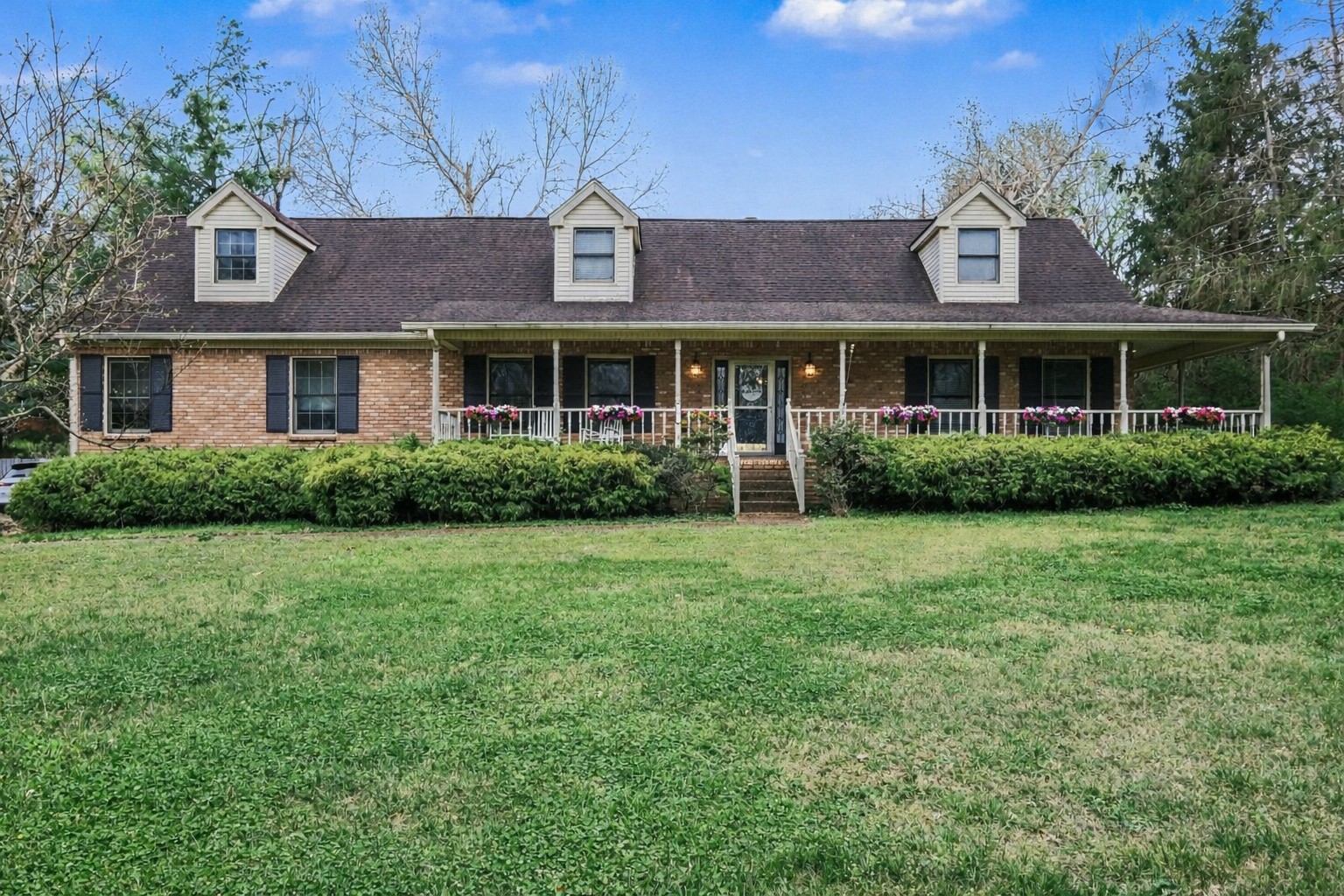 Brick home with three dormers, a flower-lined covered porch, and a broad front lawn.