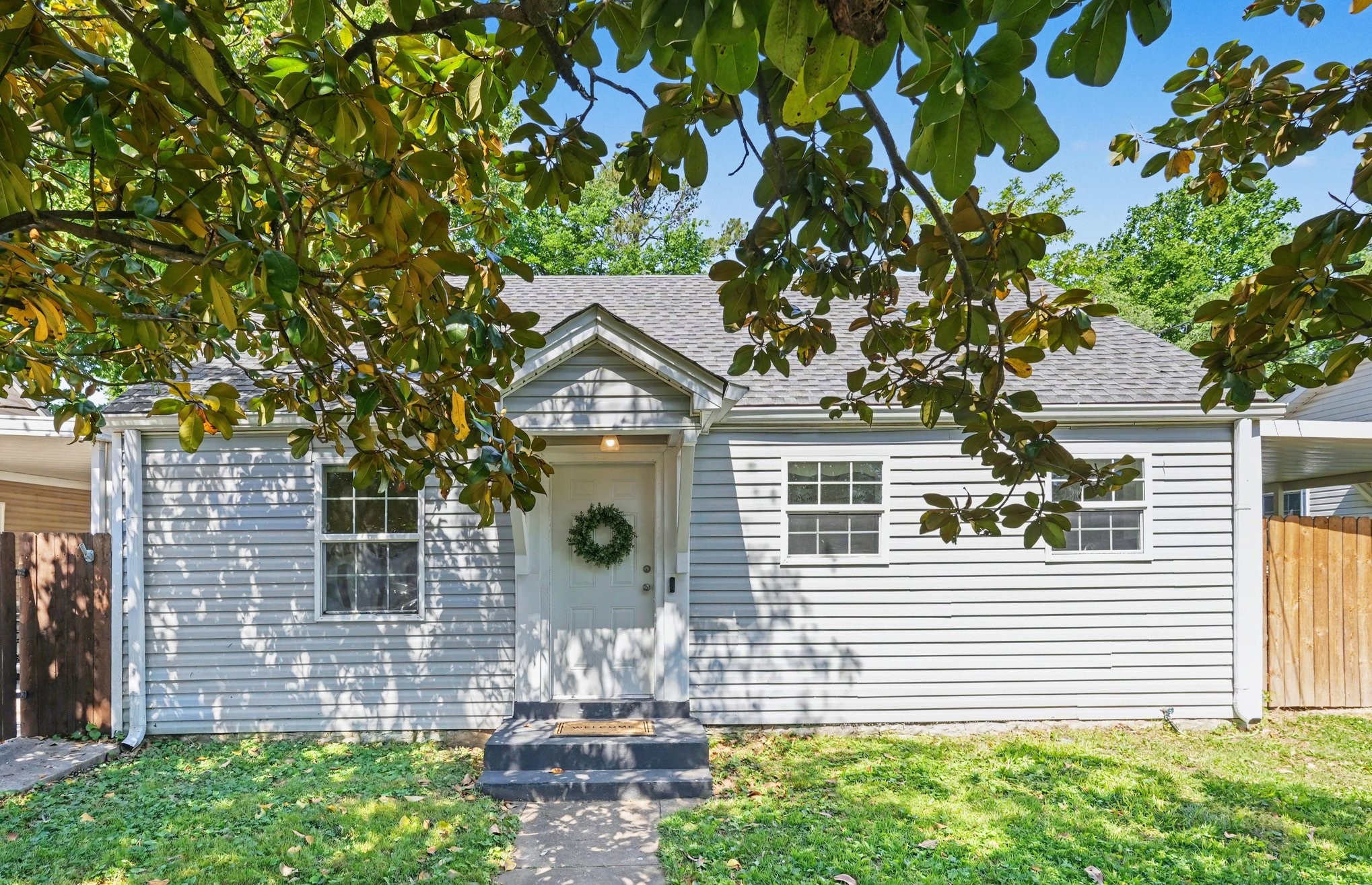 White single-story house with a gabled entry, front-door wreath, and leafy branches shading the facade.