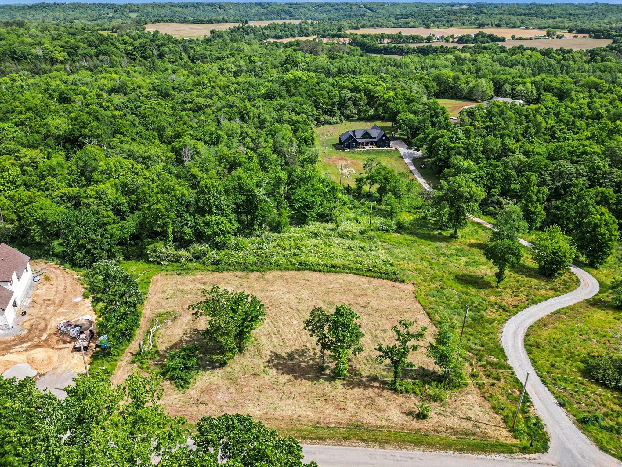 Aerial view of a cleared rural lot surrounded by woodland, with a winding drive and a house beyond.