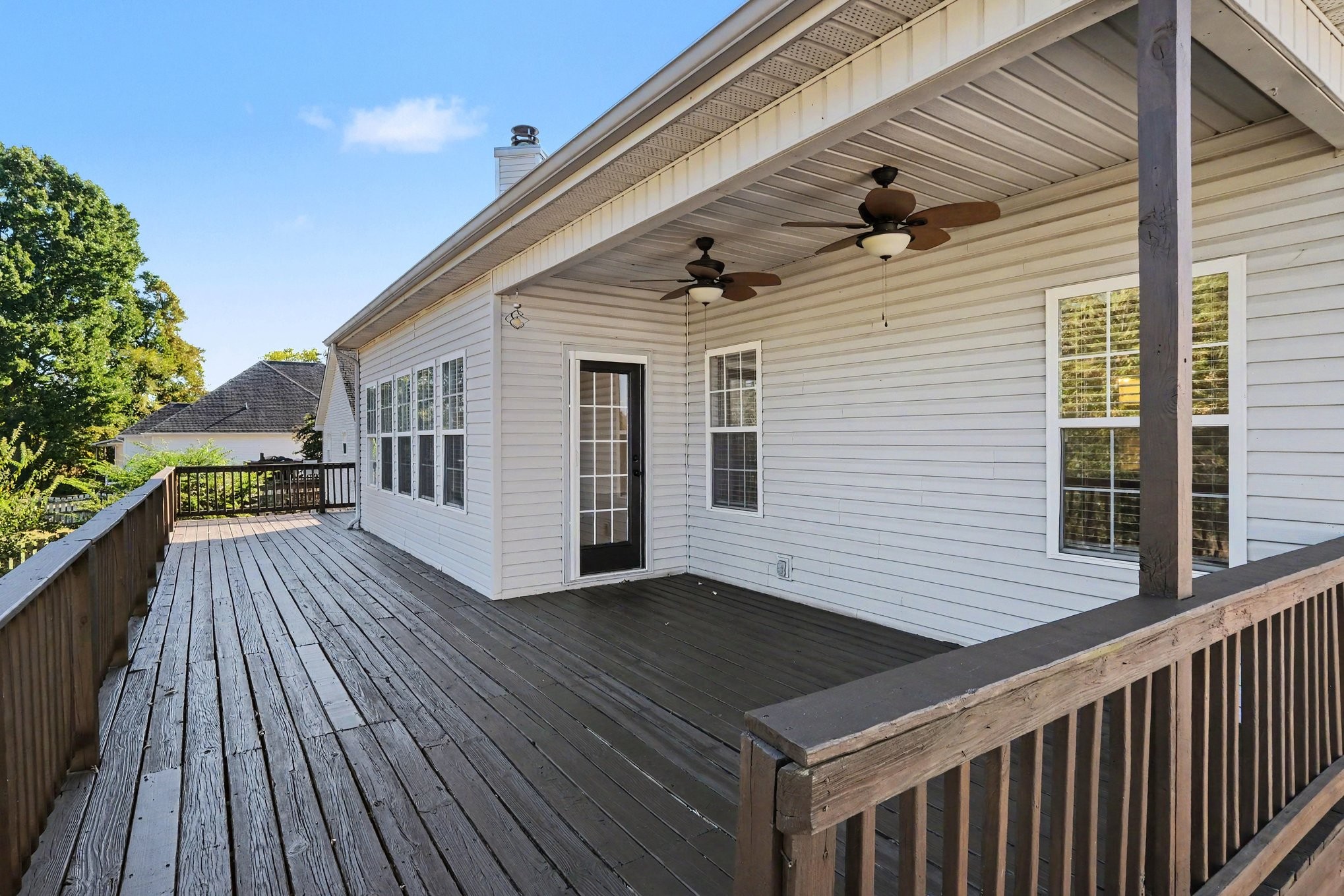 Covered wooden deck with two ceiling fans, white siding, and railings overlooking leafy trees.