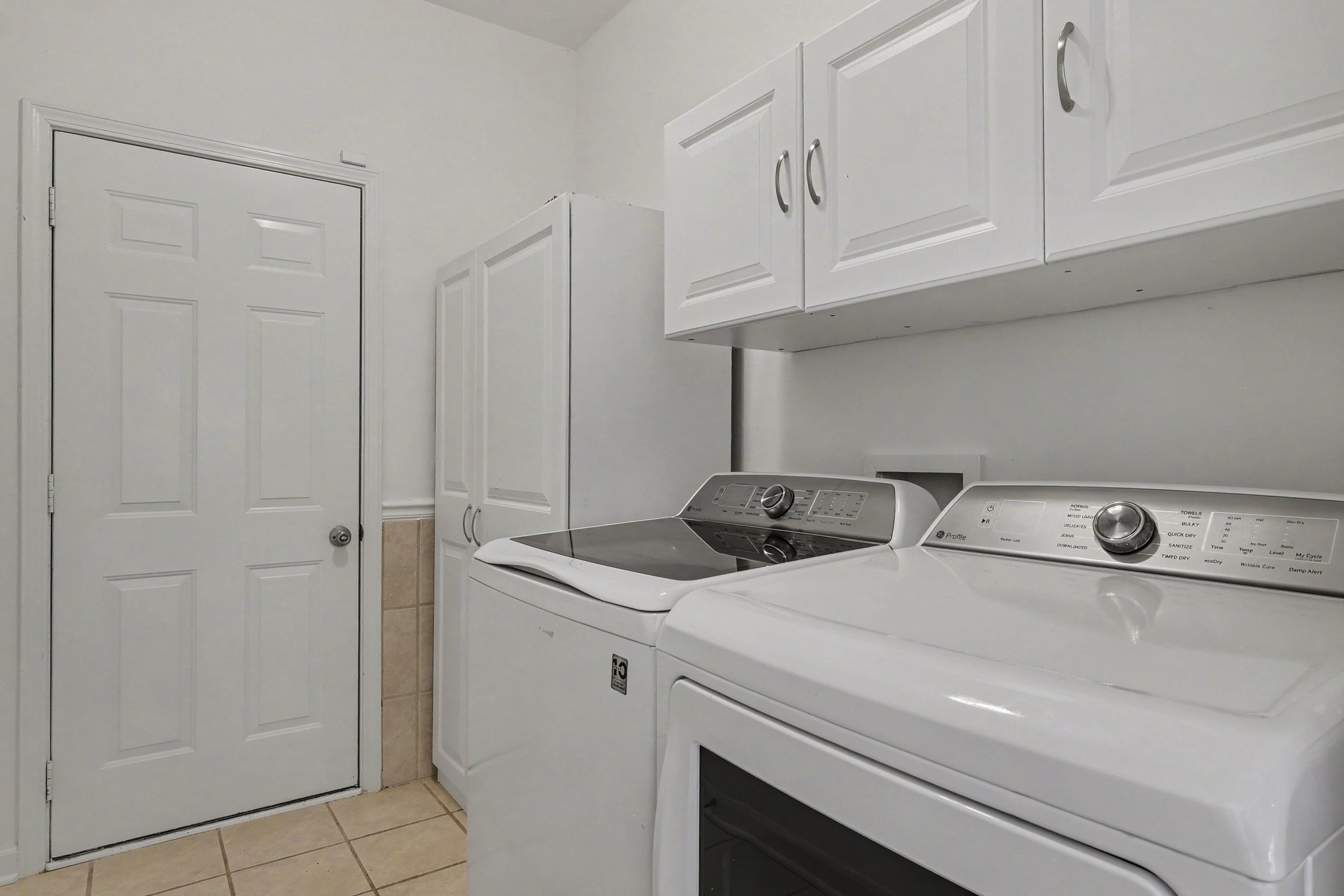 Laundry room with white washer and dryer beneath upper cabinets beside a paneled door.