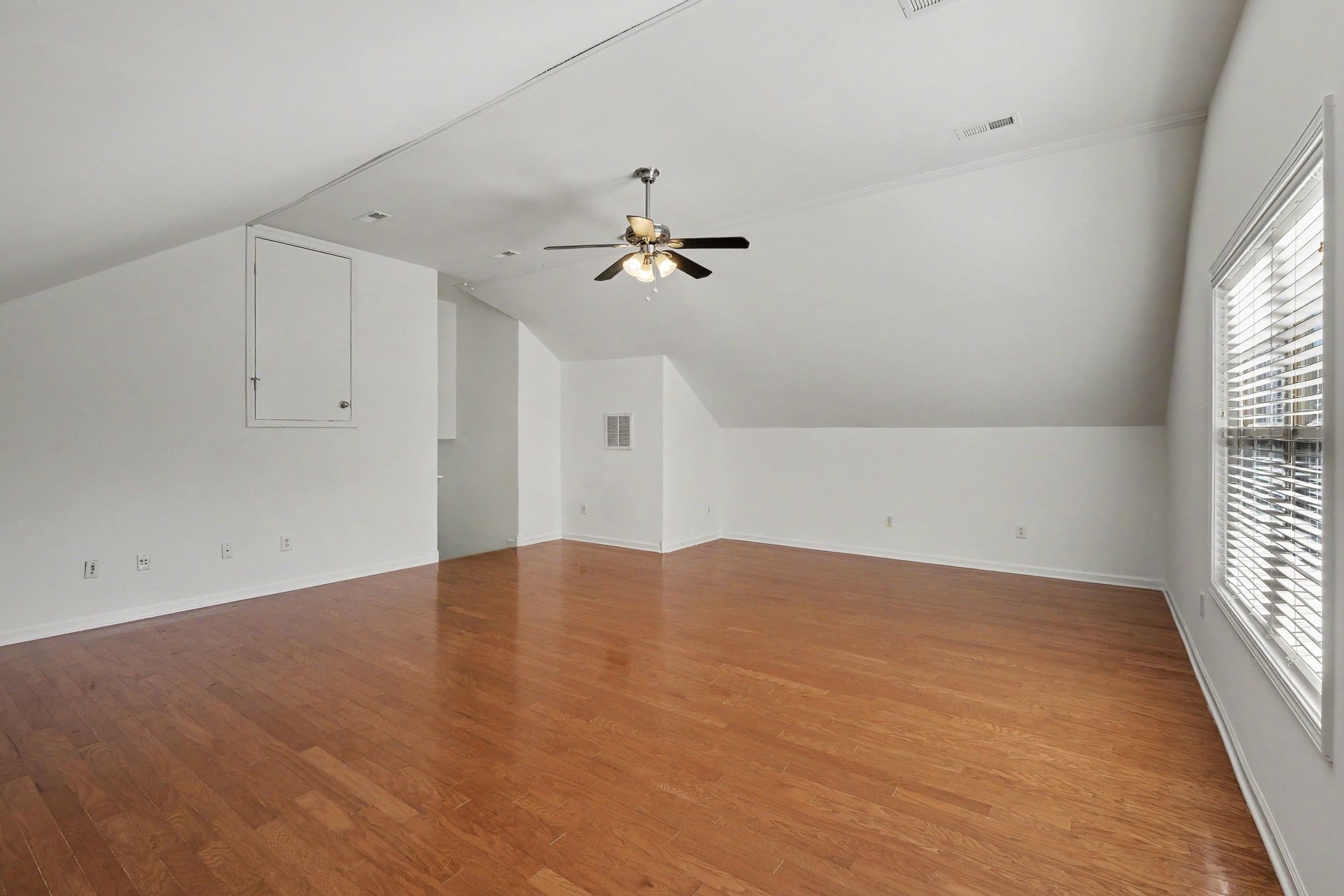 Empty upstairs room with hardwood floors, a sloped ceiling, ceiling fan, and large window with blinds.
