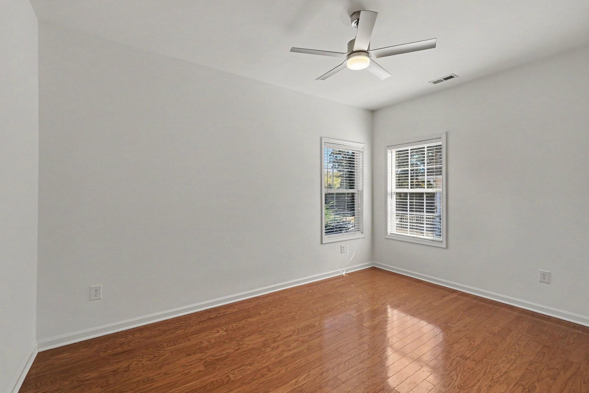 Empty room with hardwood floors, two blinded windows, and a ceiling fan.