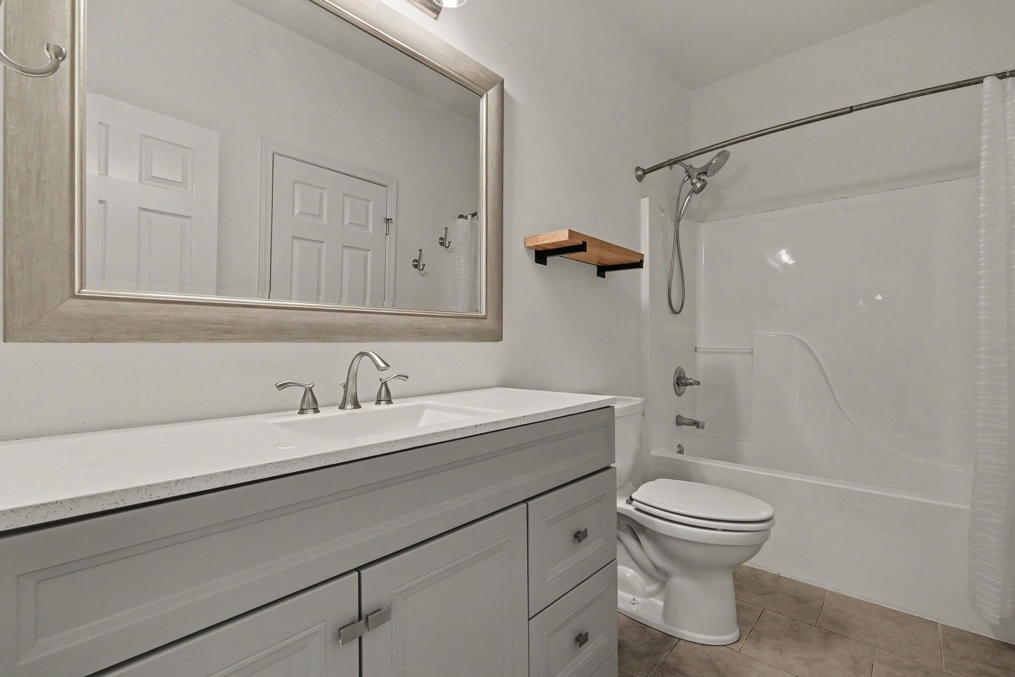 Bathroom with gray vanity and framed mirror beside a white tub-shower and toilet.