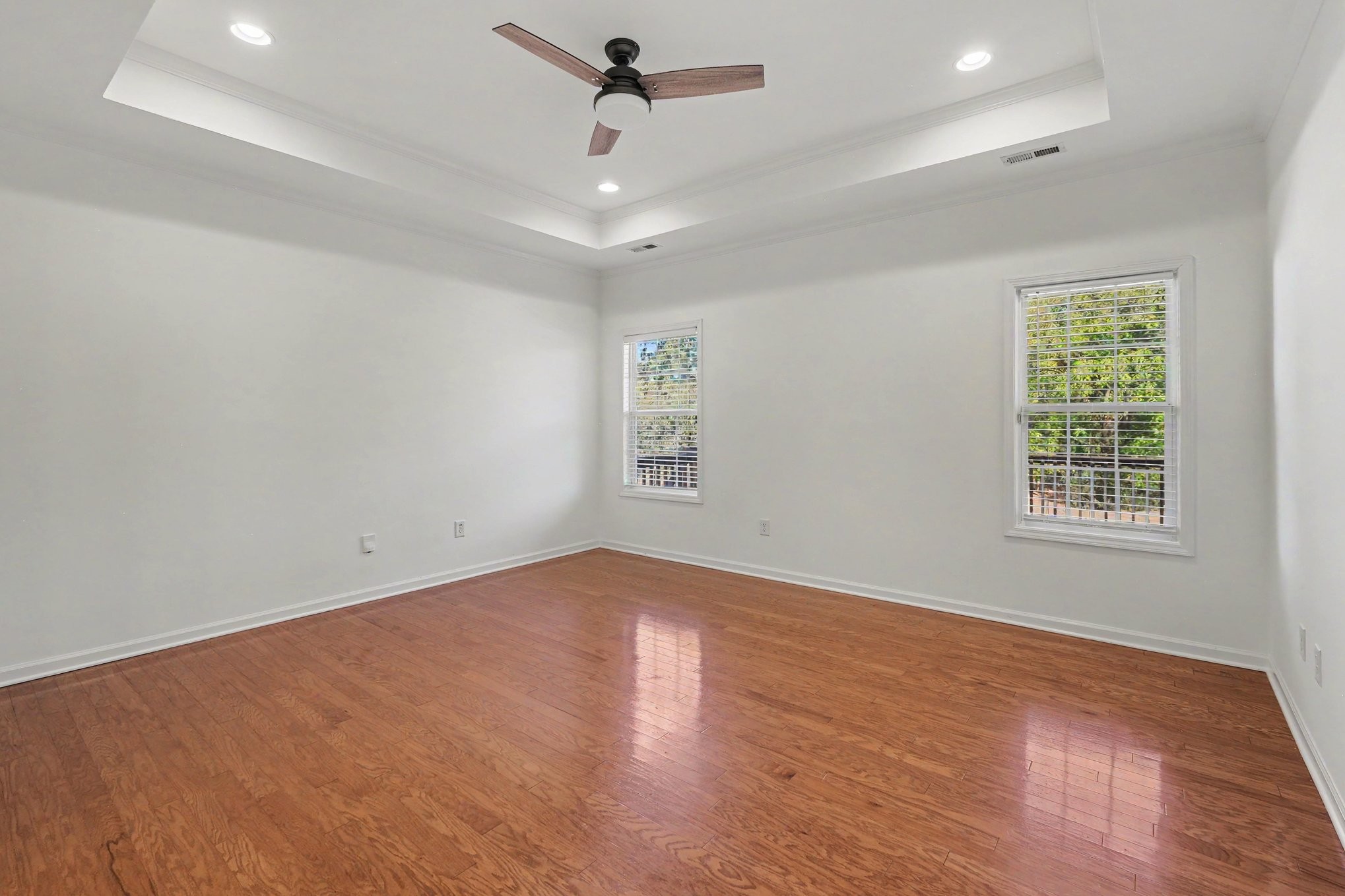 Empty room with hardwood floors, two windows, and a tray ceiling with ceiling fan.