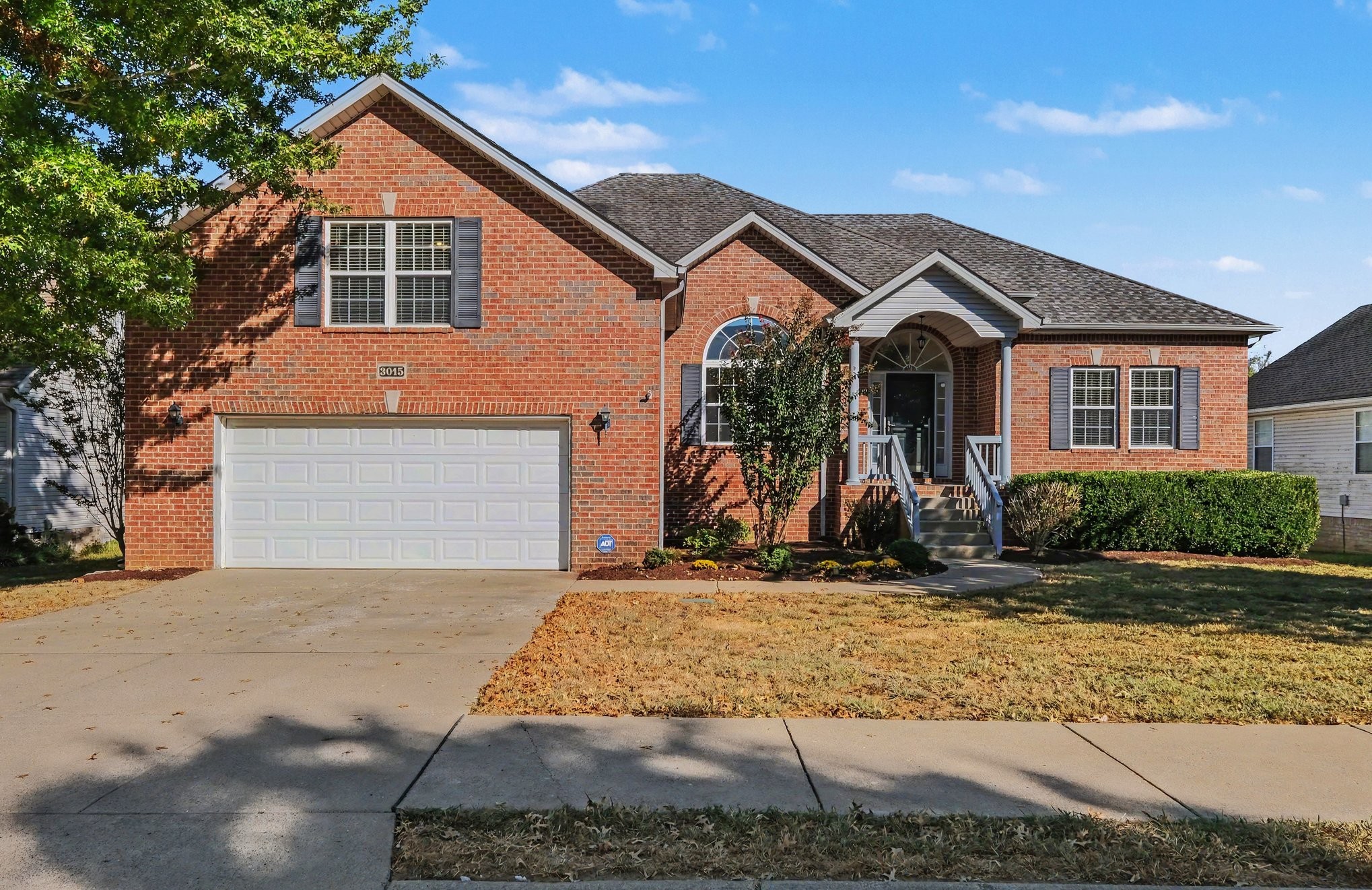 Red-brick two-story home with a double garage, covered arched entry, and autumn-brown lawn.