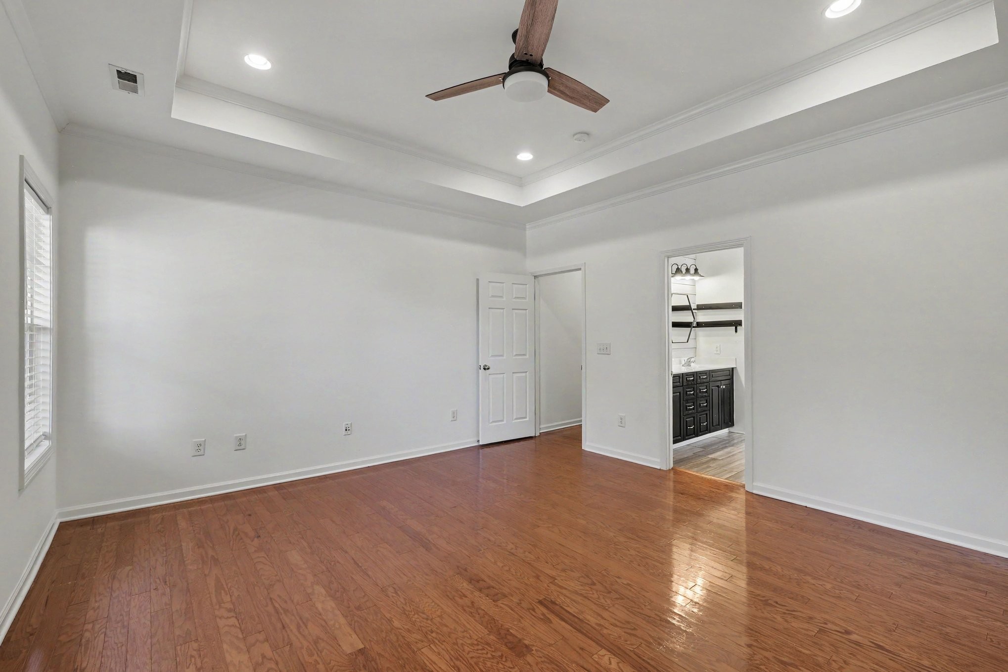 Vacant bedroom with hardwood floors, tray ceiling, ceiling fan, and an adjoining bathroom.
