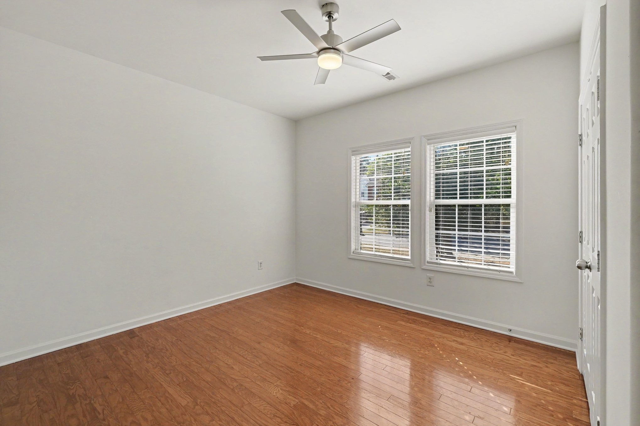 Empty bedroom with hardwood floors, a ceiling fan, and two windows with blinds.