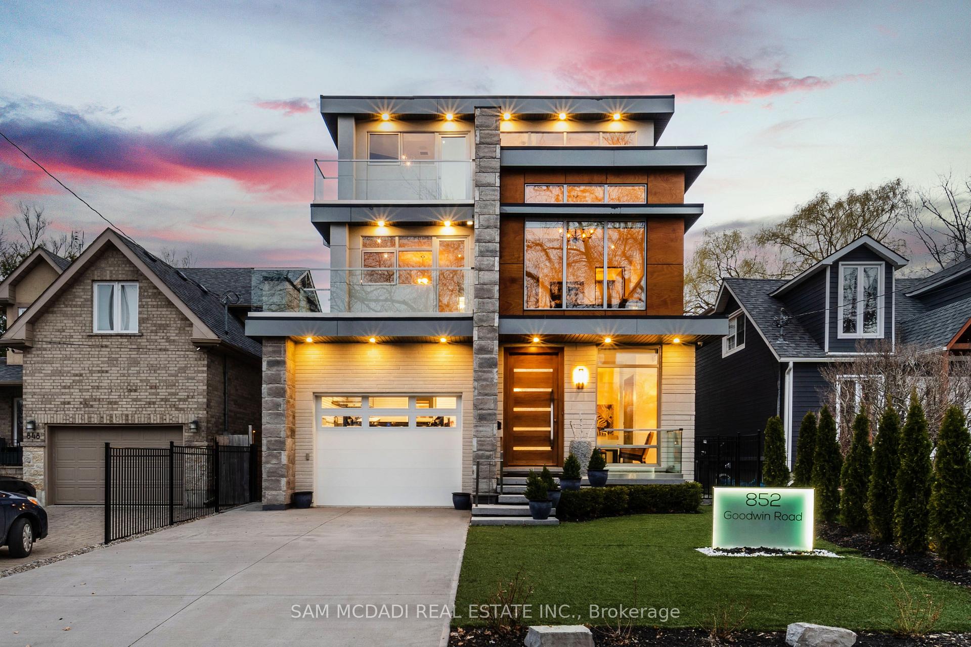 Modern three-story house with glass balconies and warm lighting at dusk, with an 852 Goodwin Road lawn sign.