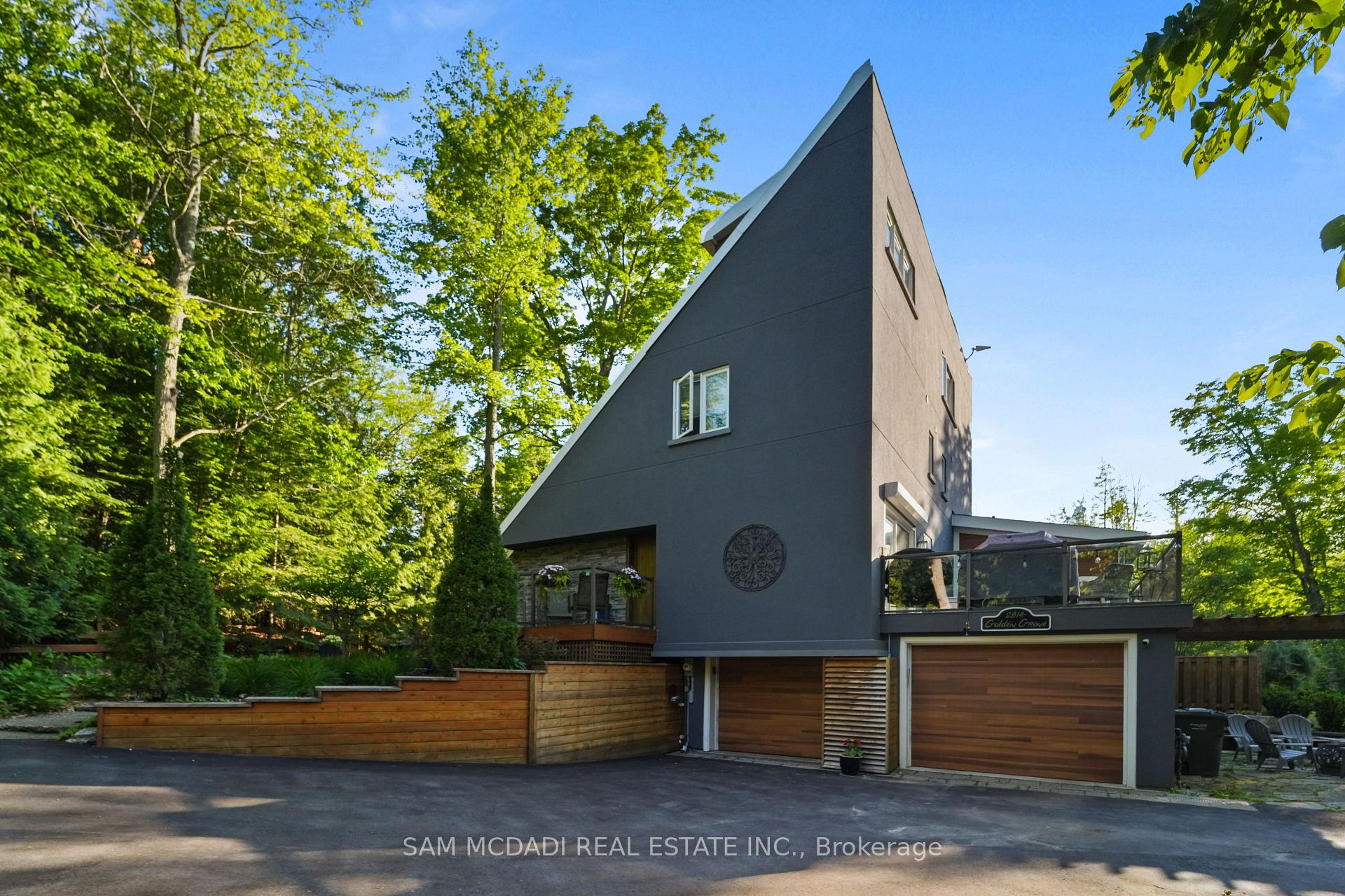 Modern gray home with a sharply angled roof, garage, and glass-railed patio among mature trees.