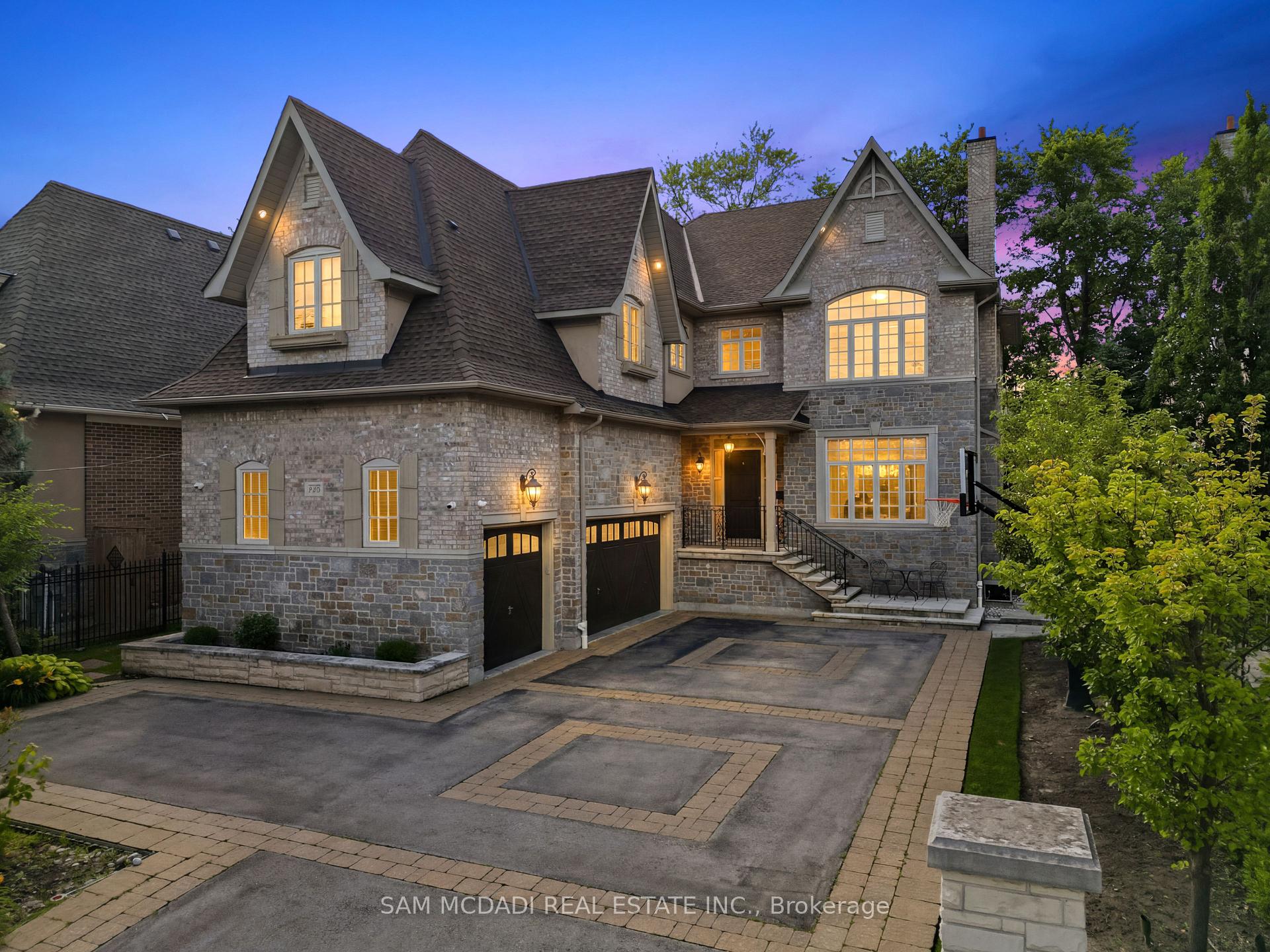 Two-story stone house with steep gables, glowing windows, double garage, and broad paved driveway at dusk.